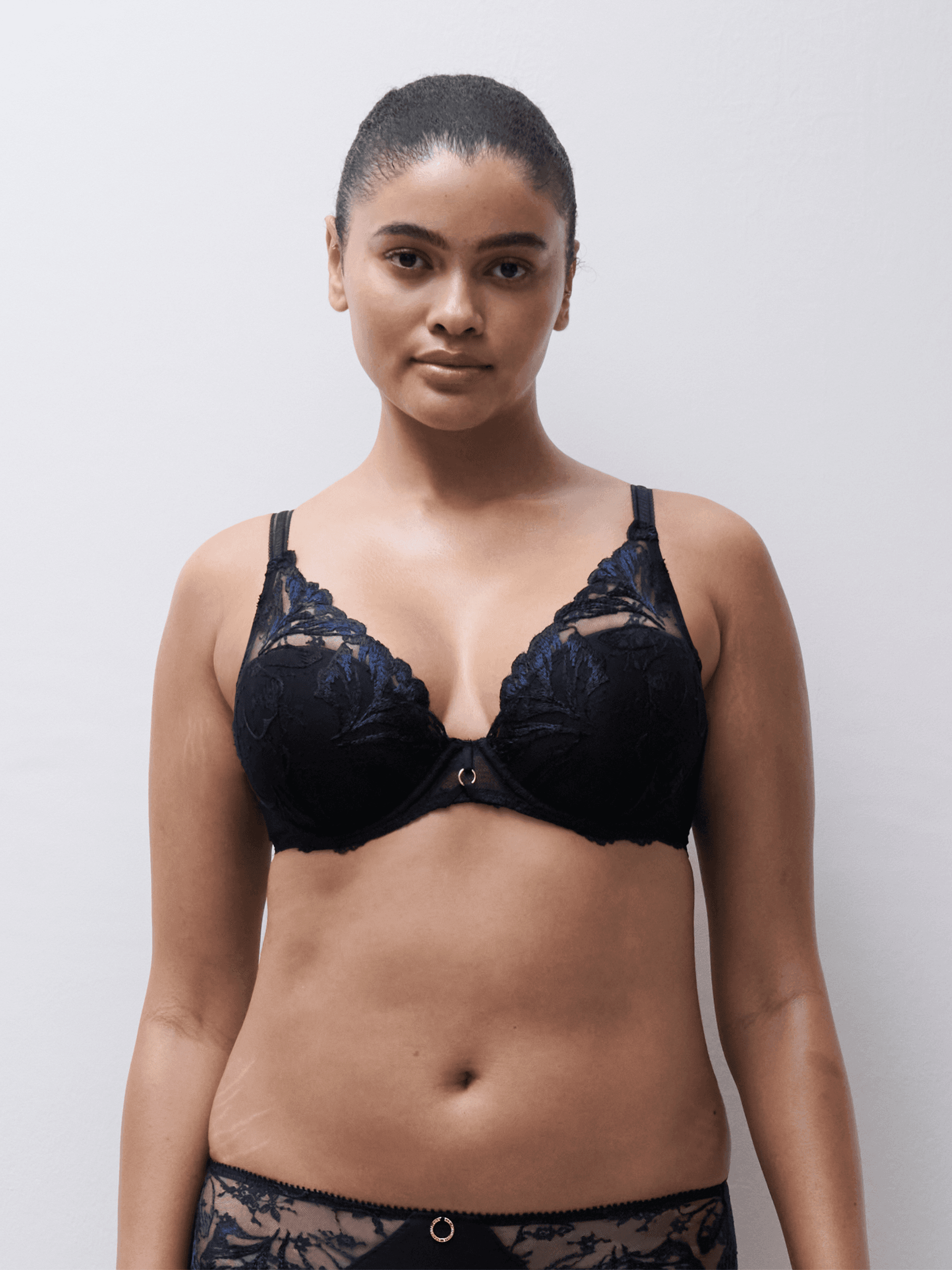 A woman stands against a plain background, wearing a black lace bra and matching underwear, with her hair tied back.