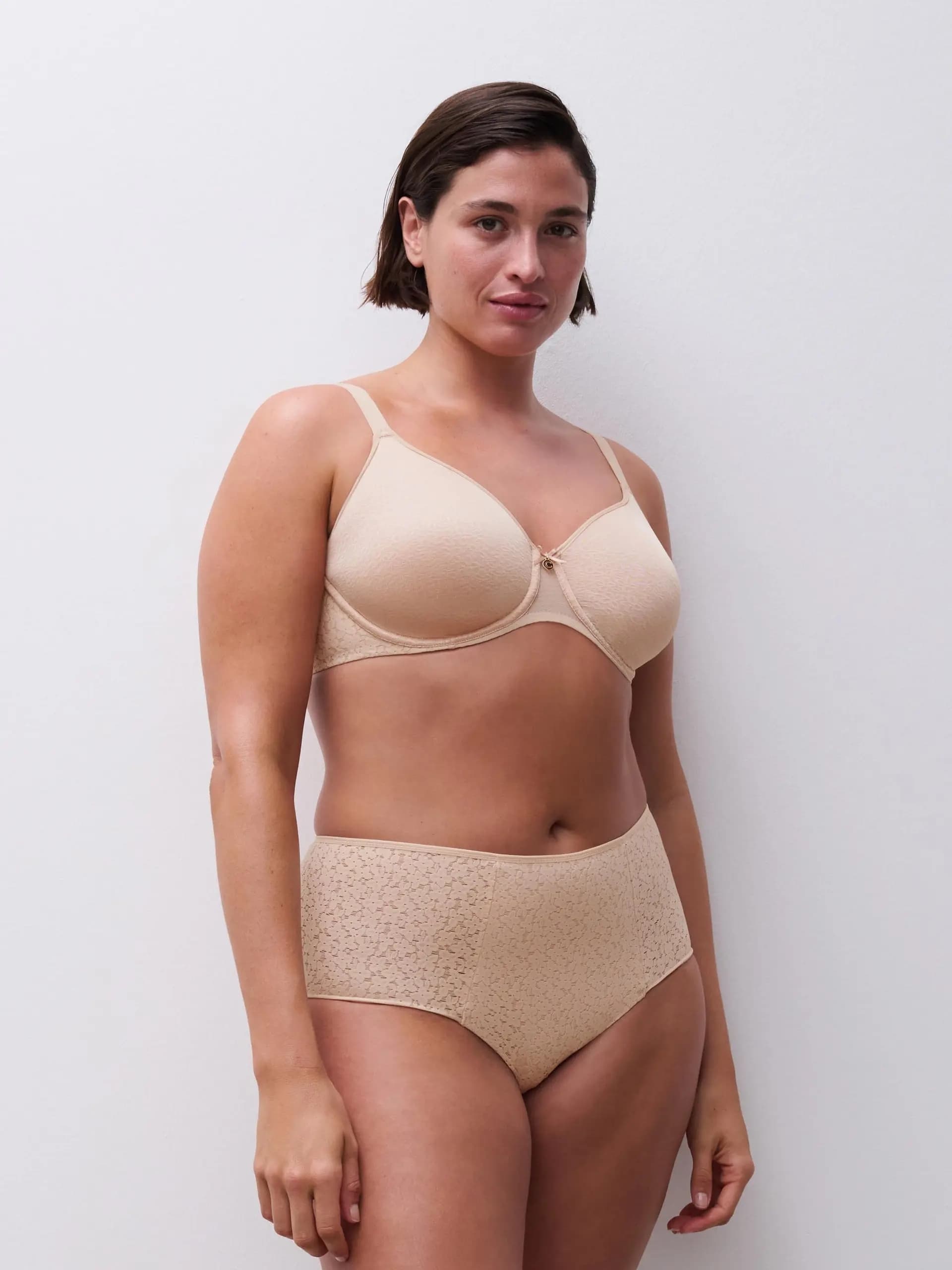 A woman in beige lingerie stands confidently against a plain white background, showcasing a bra and high-waisted panties.