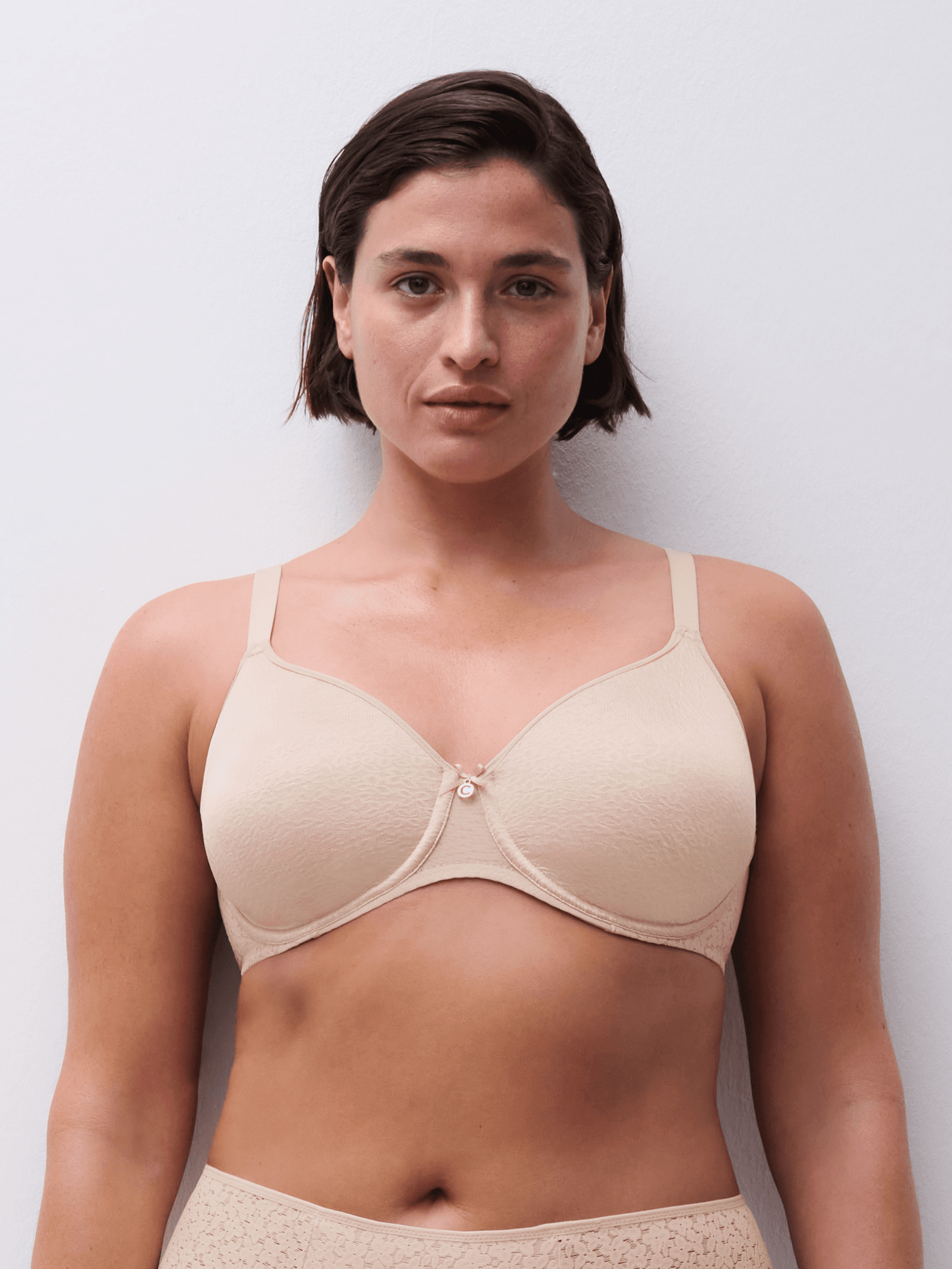 A person with short dark hair wearing a skin-toned bra, standing against a plain white background.