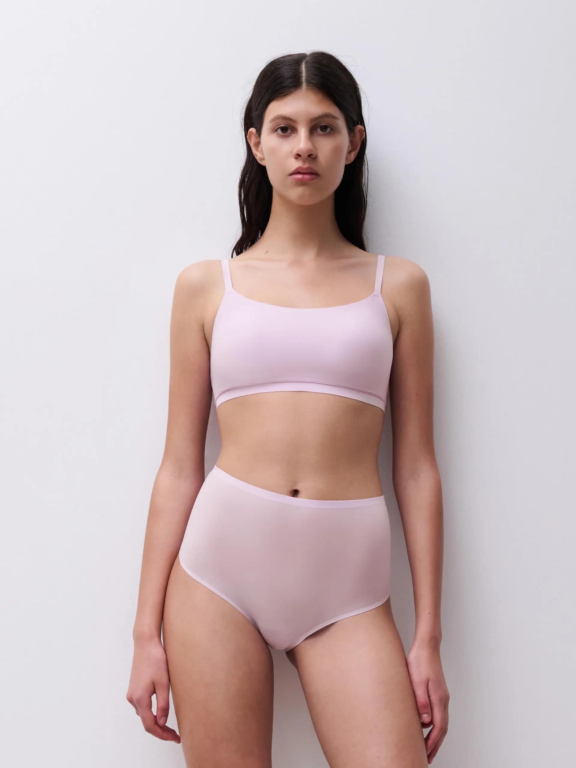 Woman in light pink underwear stands against a white background, looking directly at the camera.