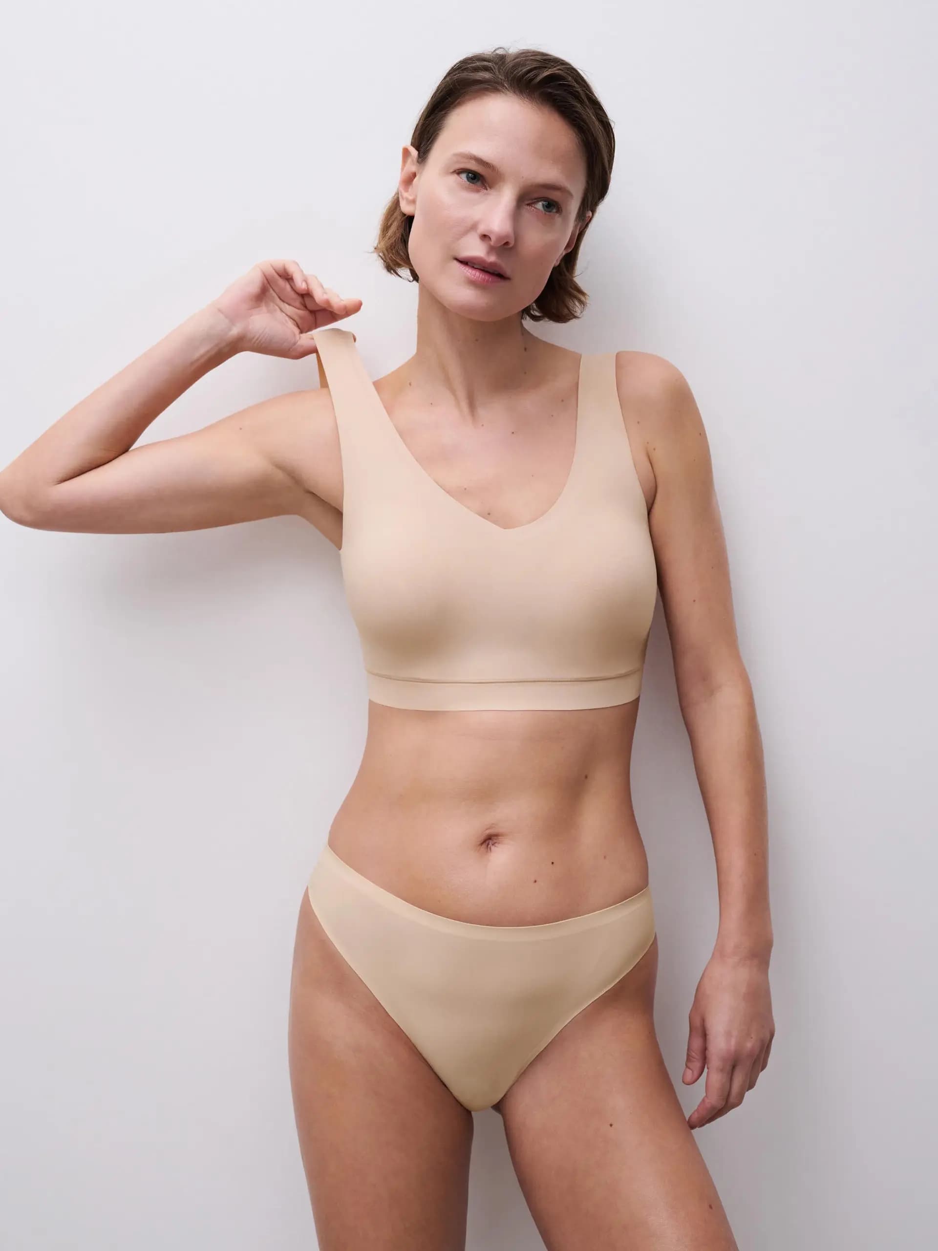 Person wearing a seamless, beige bra and panties set, standing against a plain white background, looking thoughtful.