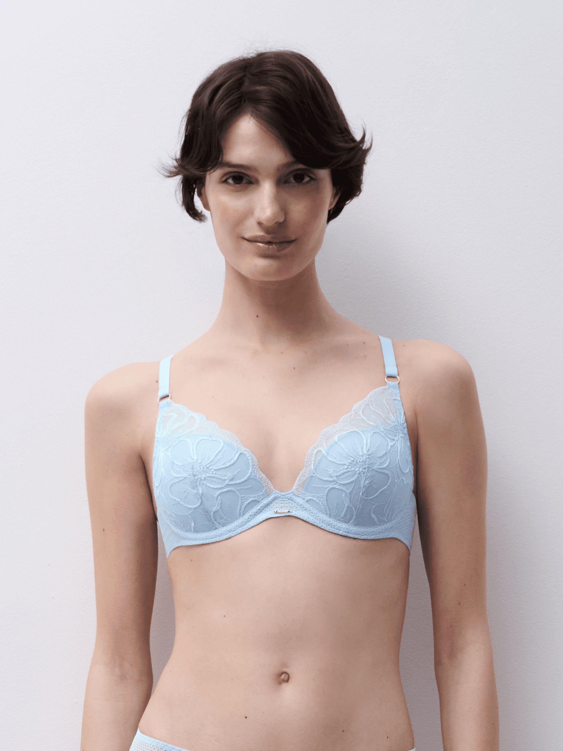 A person with short hair wearing a light blue bra with floral lace design, standing against a plain background.