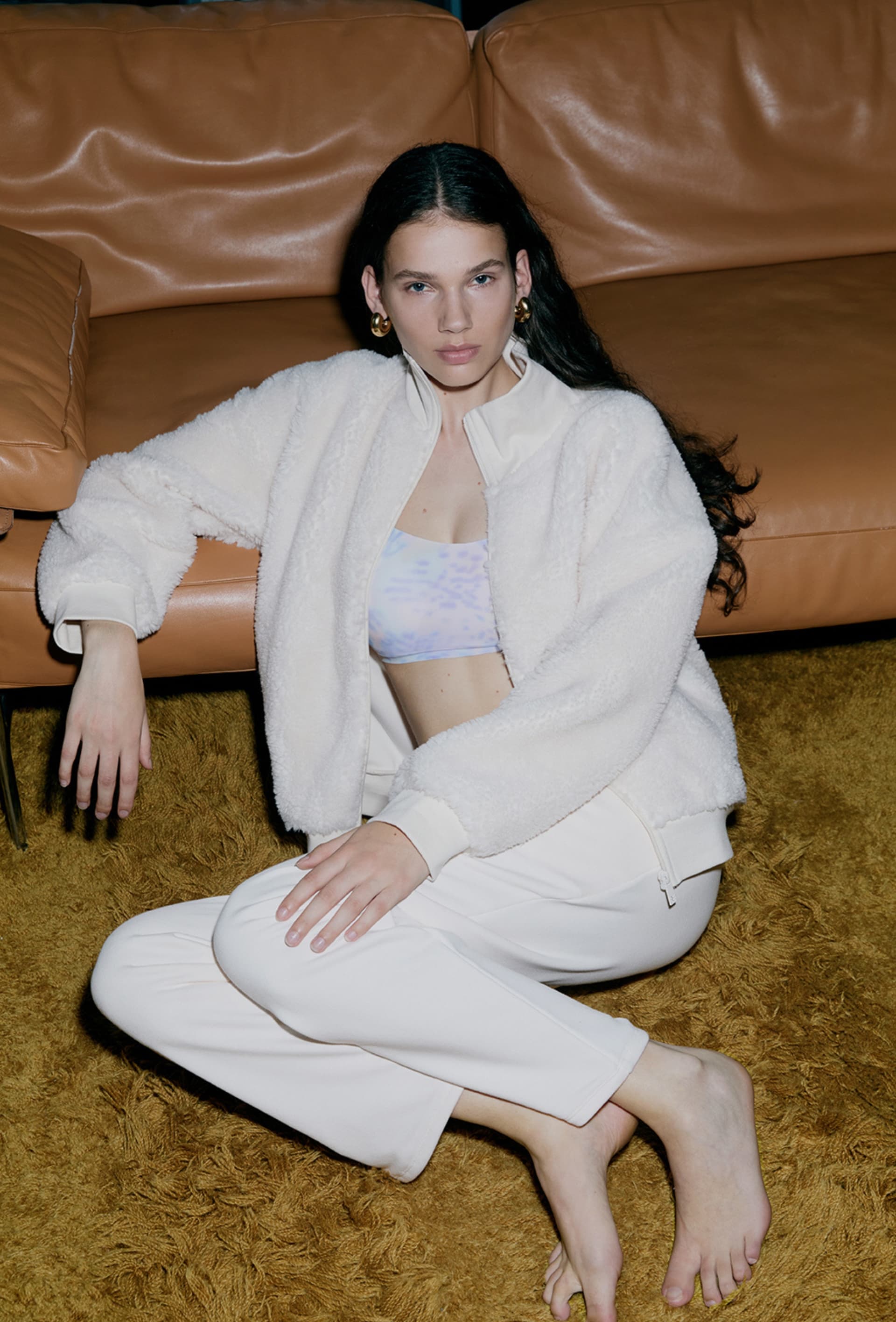 Woman in white fuzzy jacket and leggings sits barefoot on a shag carpet, leaning against a tan leather sofa, with a neutral expression.