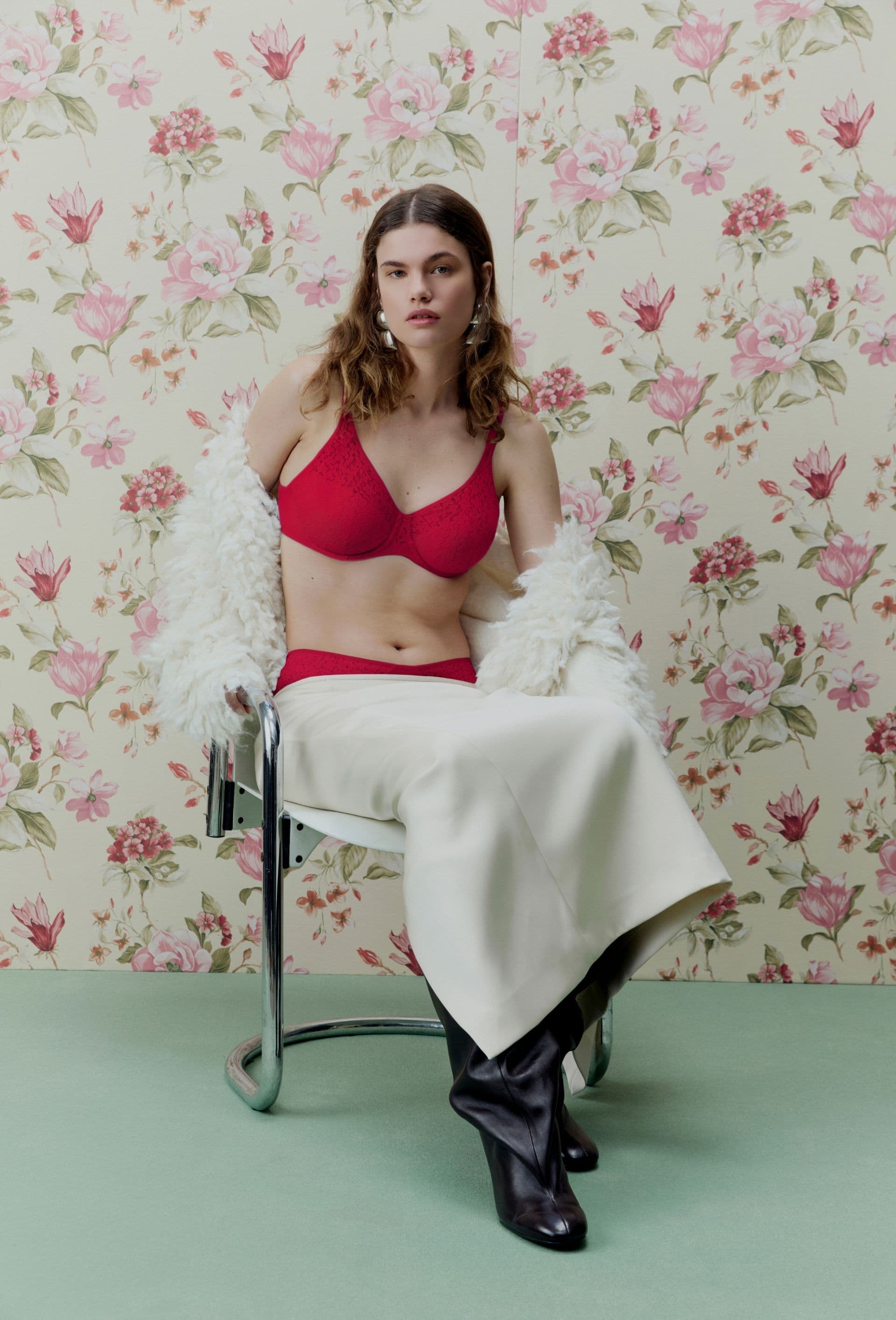 Person in a red bra and white skirt sits on a chair, draped in a fluffy white shawl, with floral wallpaper in the background.