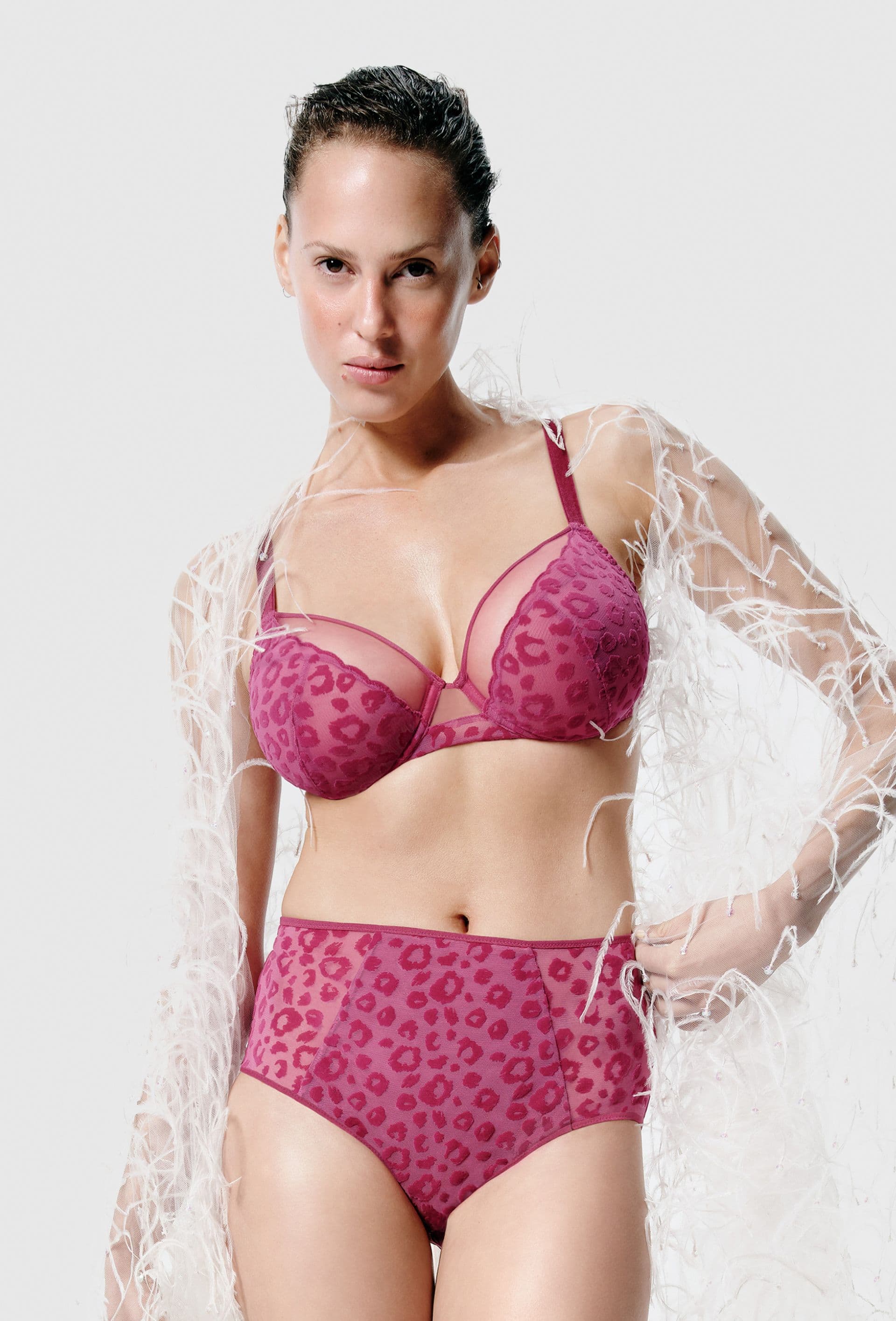 Person wearing pink leopard print lingerie with sheer detailing, draped in a feathered shawl, against a plain white background.