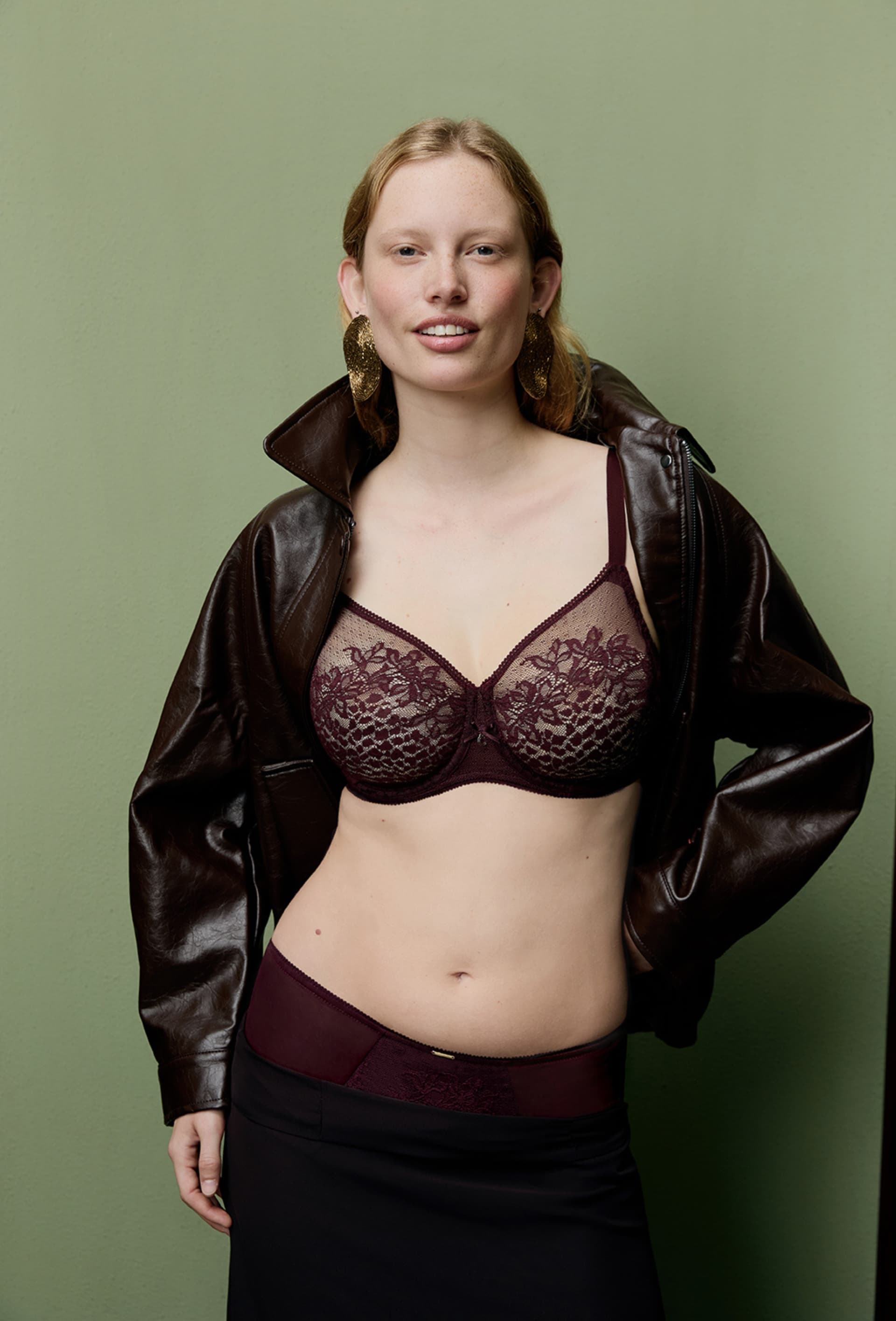 Woman in a maroon lace bra, leather jacket, and earrings, poses confidently with a smile against a green background.