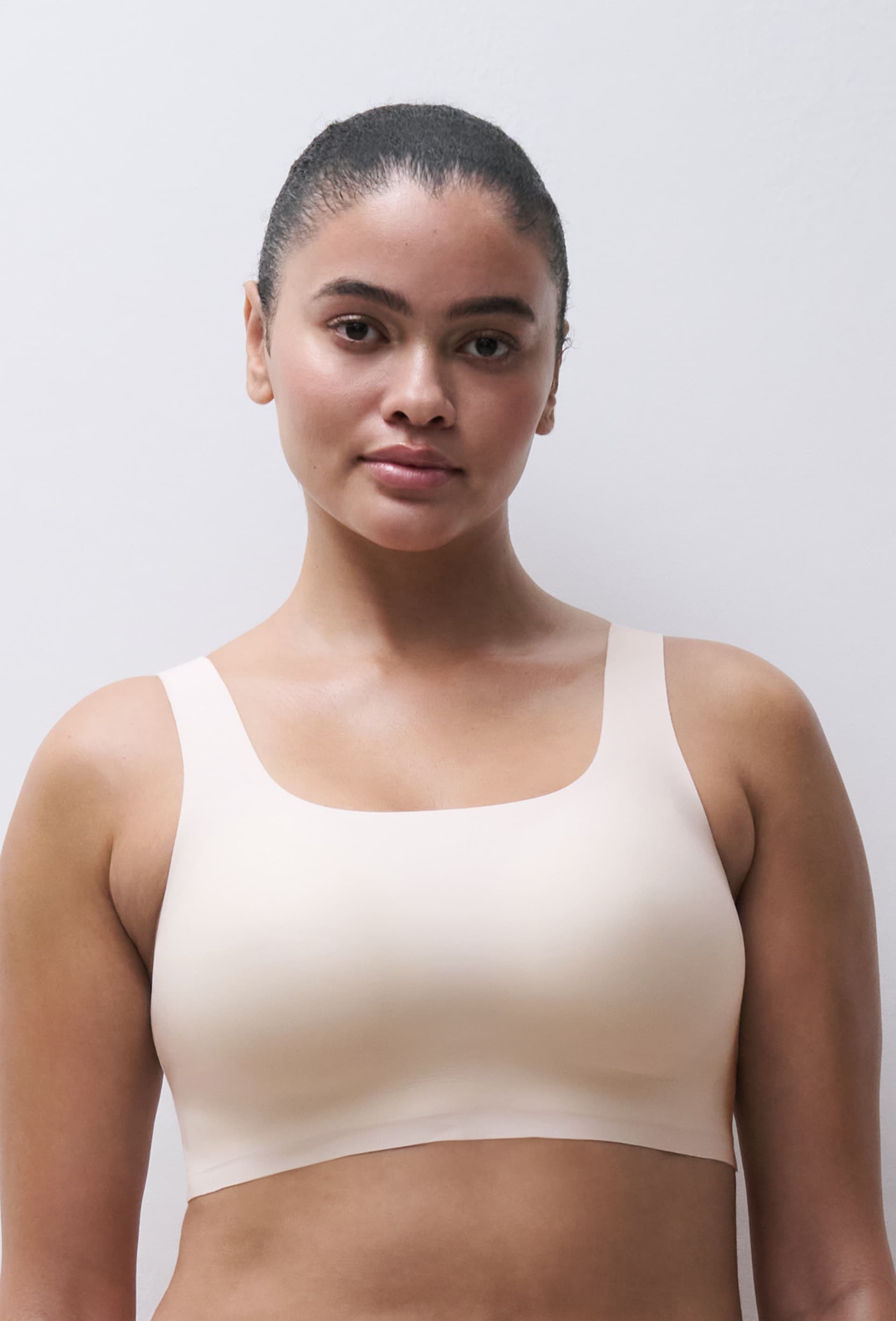 A person with tied-back hair wearing a light bra stands against a plain white background.