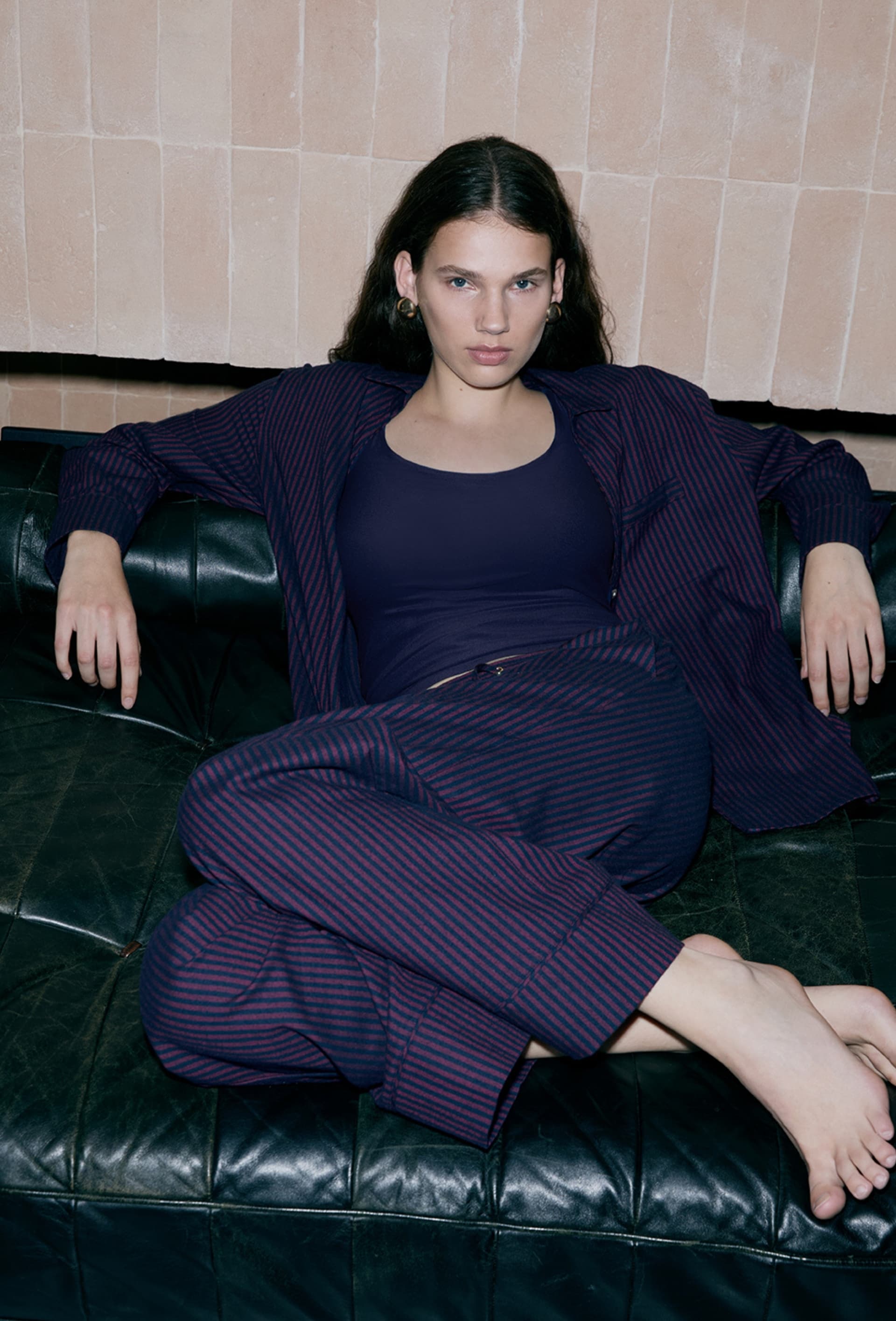 Person in a navy striped outfit lounges on a black leather couch, against a tiled wall, exuding a relaxed yet confident demeanor.