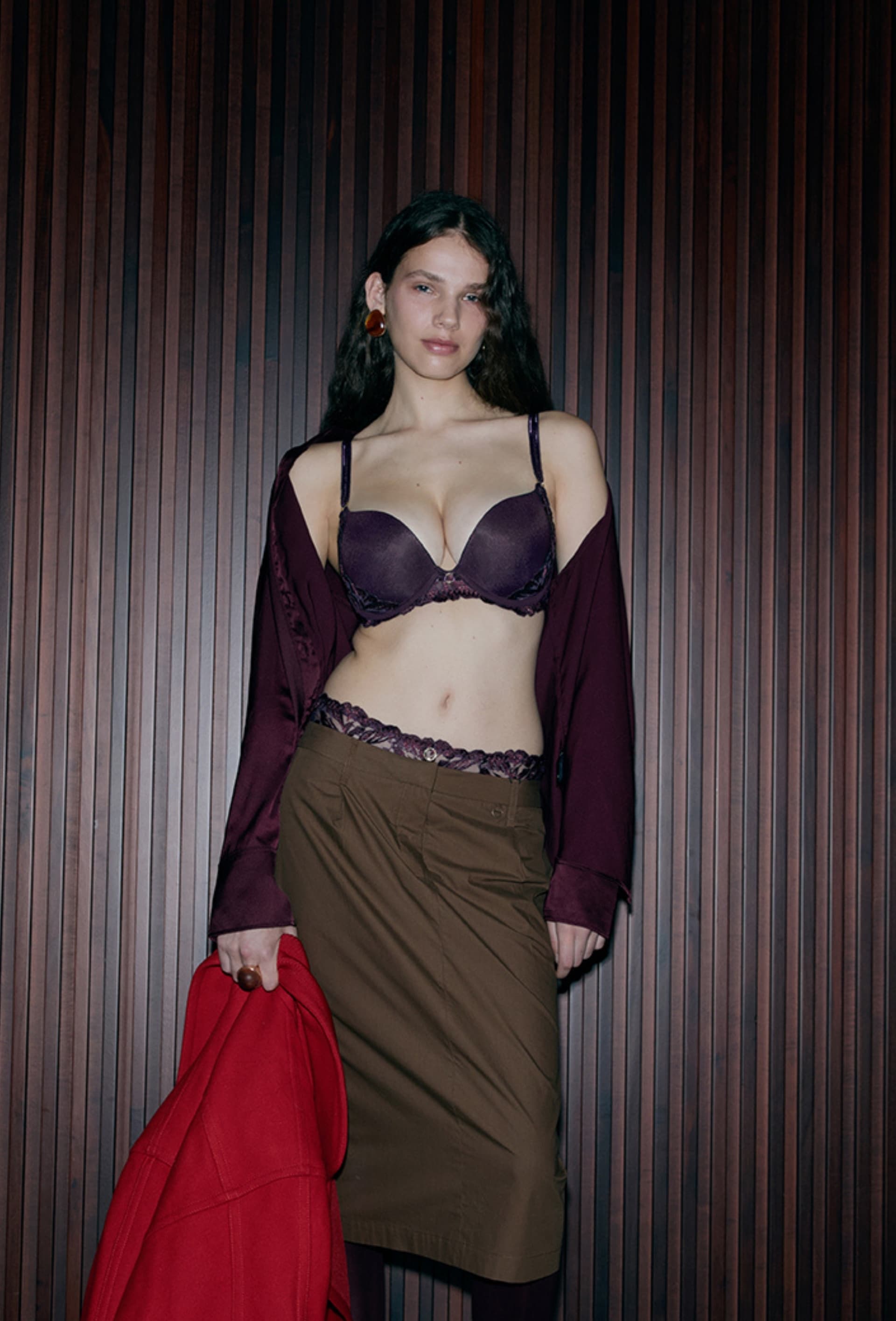 A woman in a purple lingerie set with a brown skirt and maroon shirt draped off her shoulders, holding a red coat, stands against a wooden backdrop.