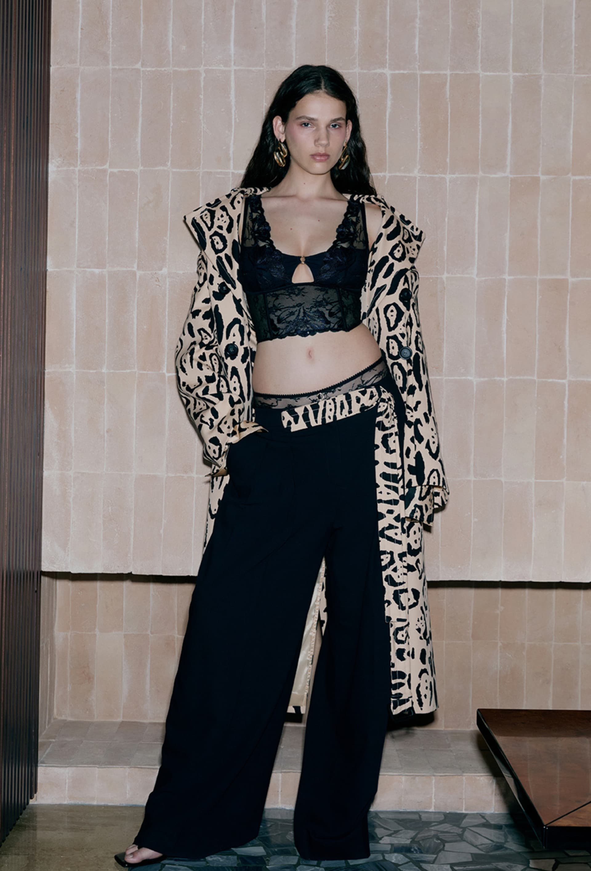Woman in leopard print coat and lace bra, black wide-leg pants, standing by a tiled wall.
