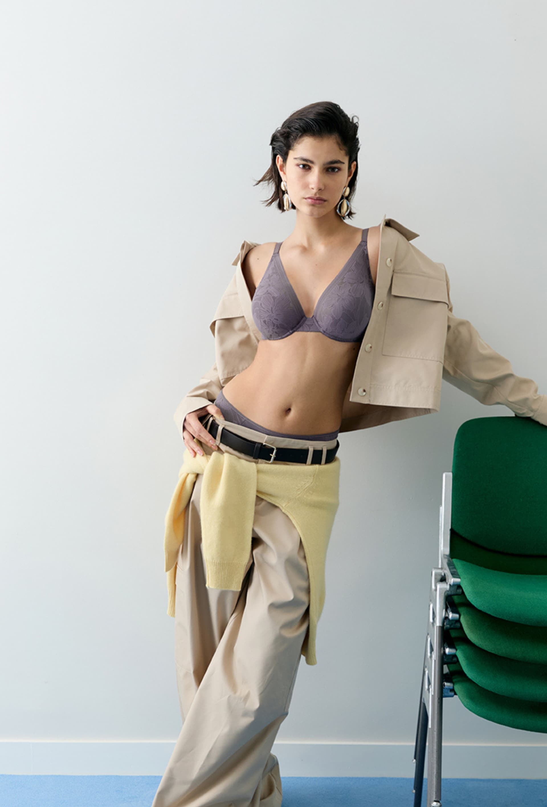 Woman in a purple bra and beige outfit, leaning against a wall near stacked green chairs.