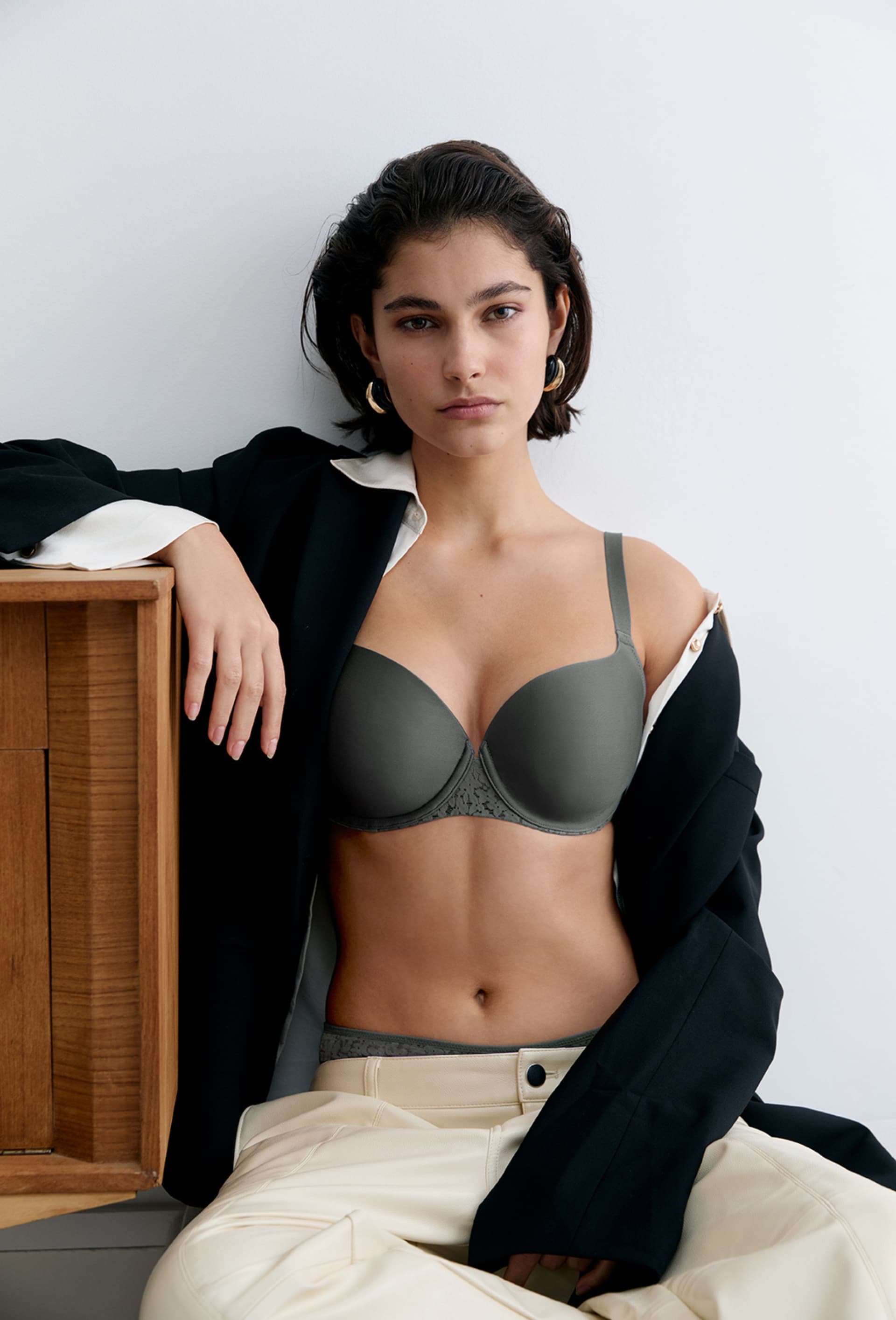 Woman in a gray bra, black blazer, and cream pants, leaning against a wooden cabinet against a white wall.