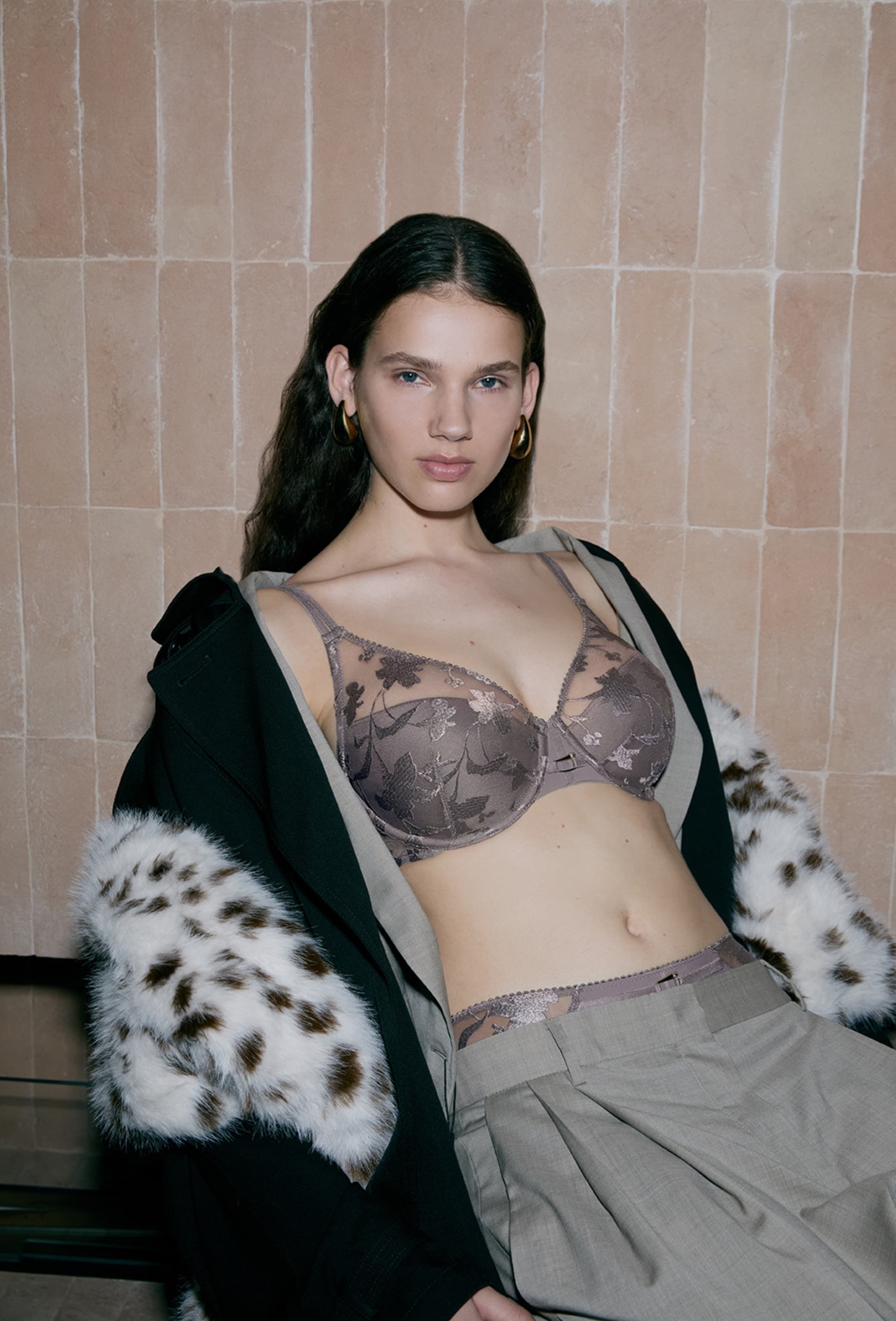 Model wearing a lace bra, gray pants, and a coat with fur lining. She poses confidently against a tiled wall.