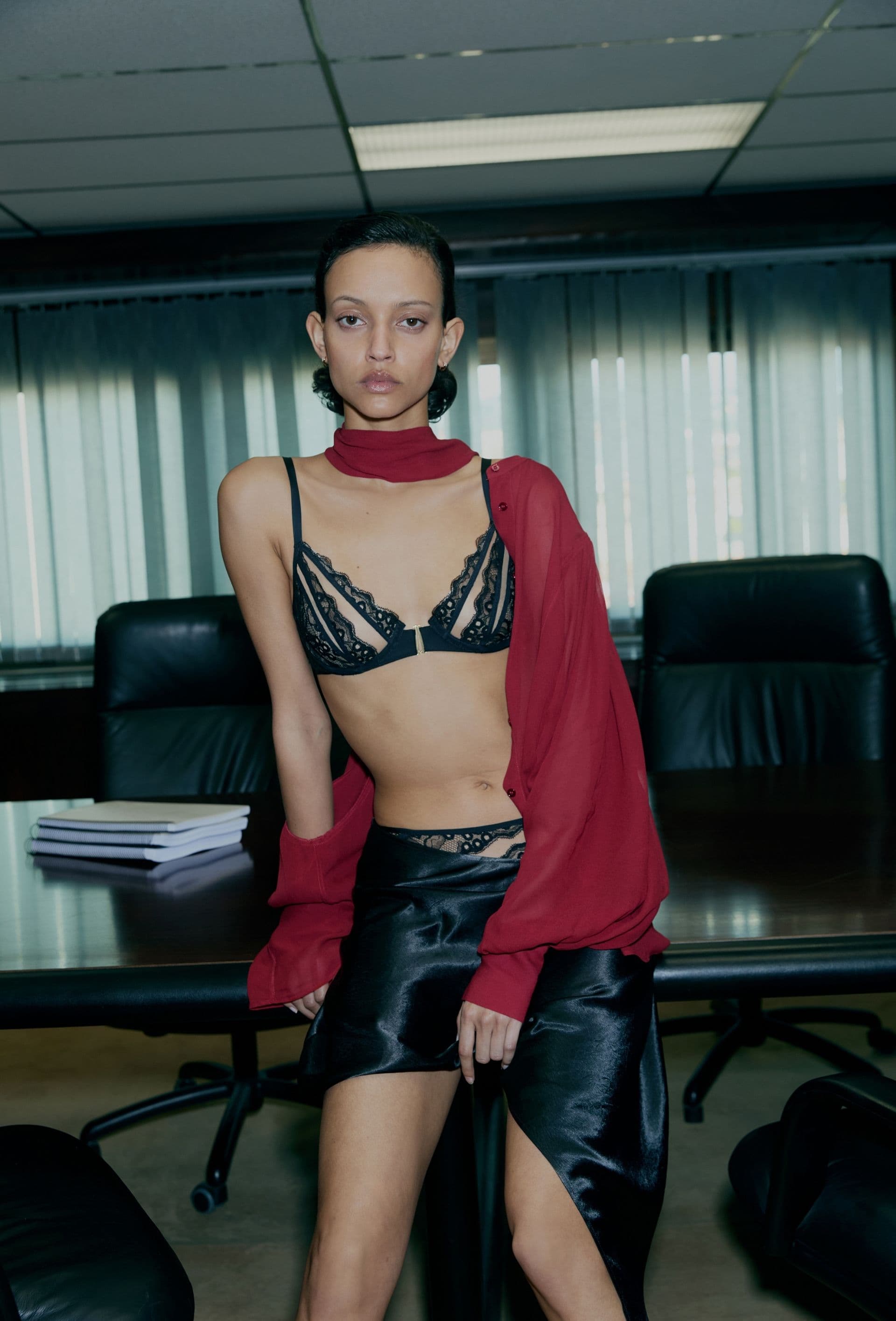 Person in a black lace bra and skirt, draped in a red scarf, stands in a dimly lit office with large windows and a wooden table.