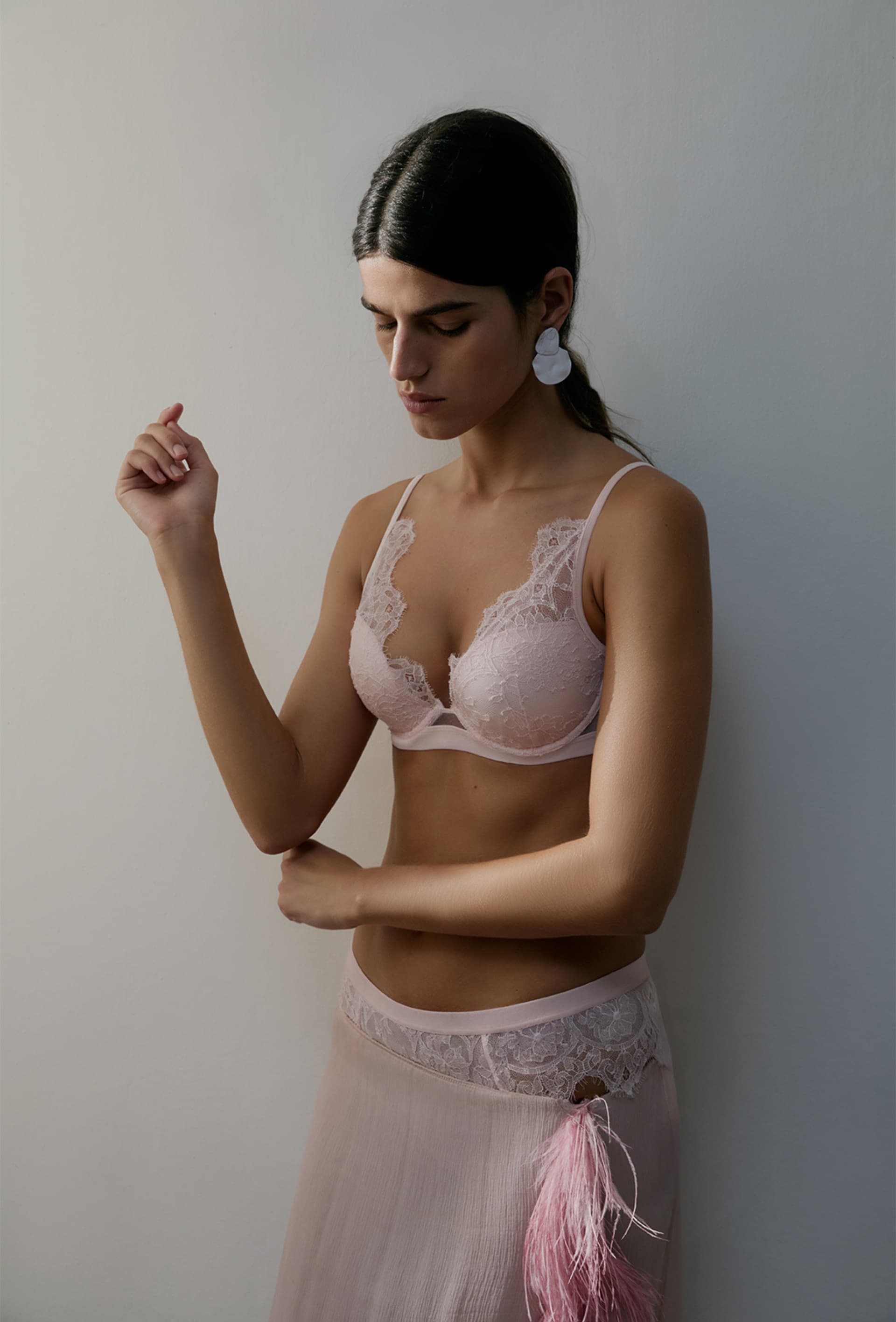 Woman in pink lace lingerie with large white earrings, standing against a white wall, looking down thoughtfully.