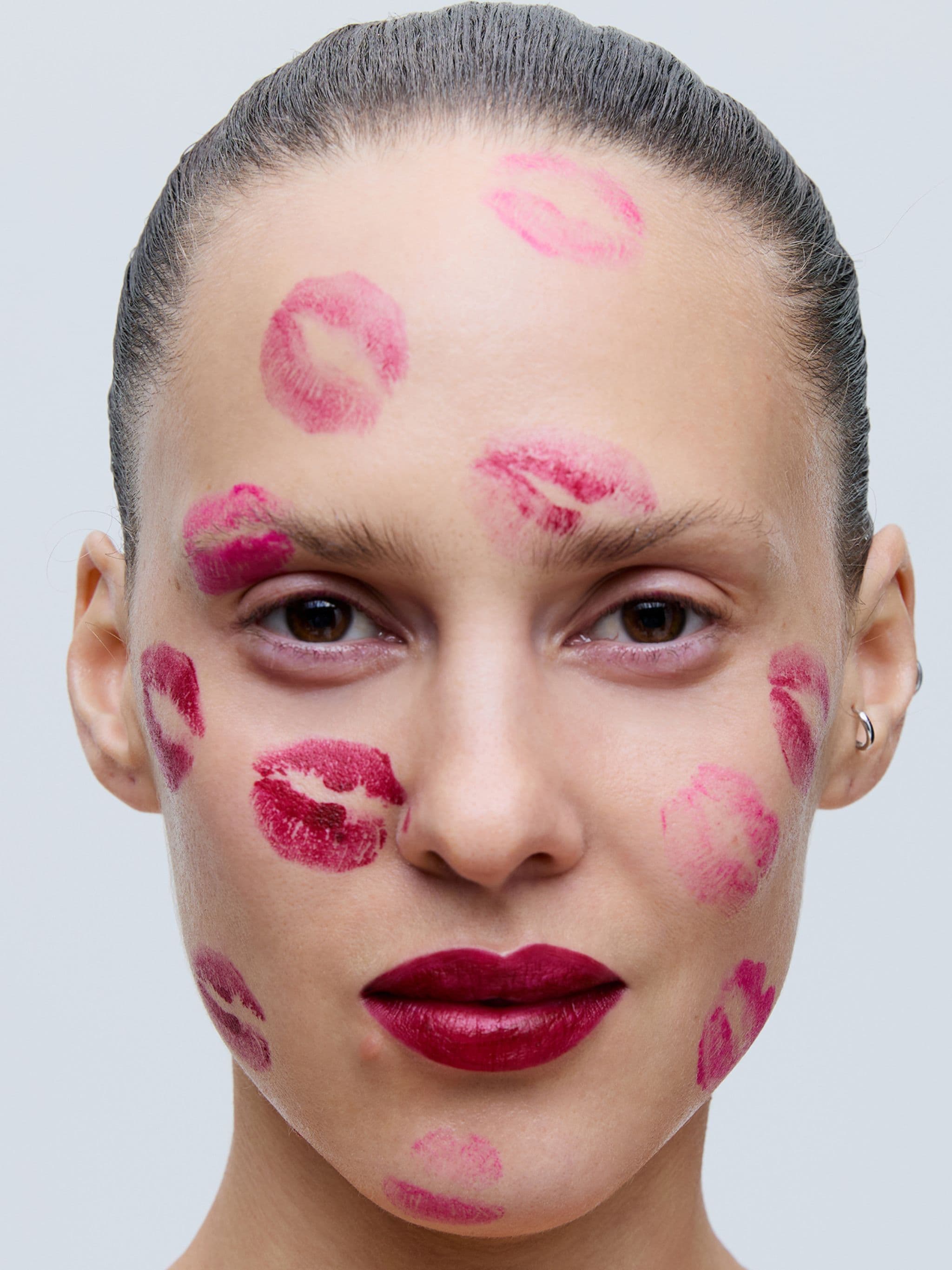 A person with sleek, pulled-back hair has red lipstick kiss marks on their face against a light gray background.