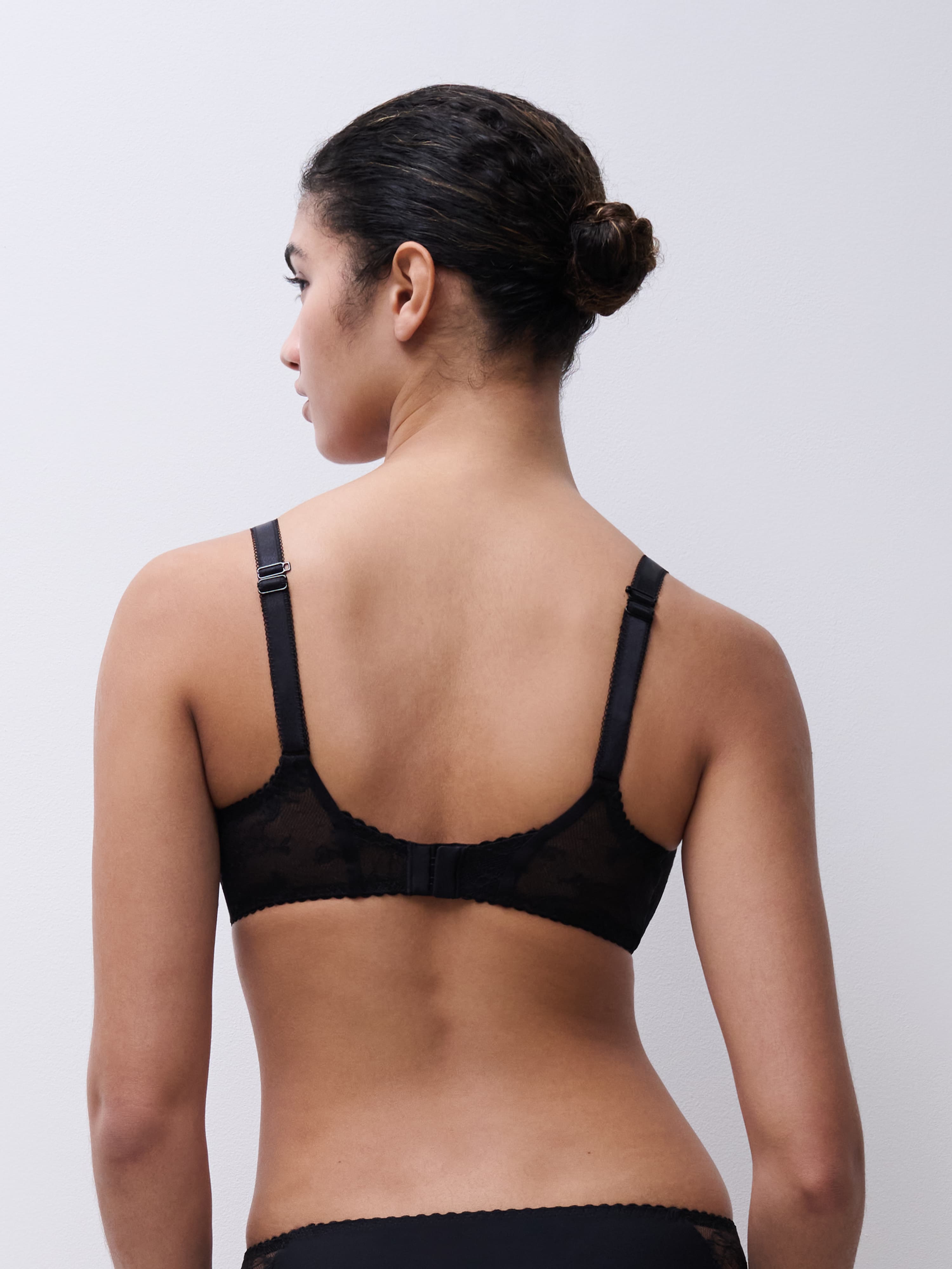 Person with short, dark hair in a bun, wearing a black lace bra and black bottoms, viewed from the back against a plain background.