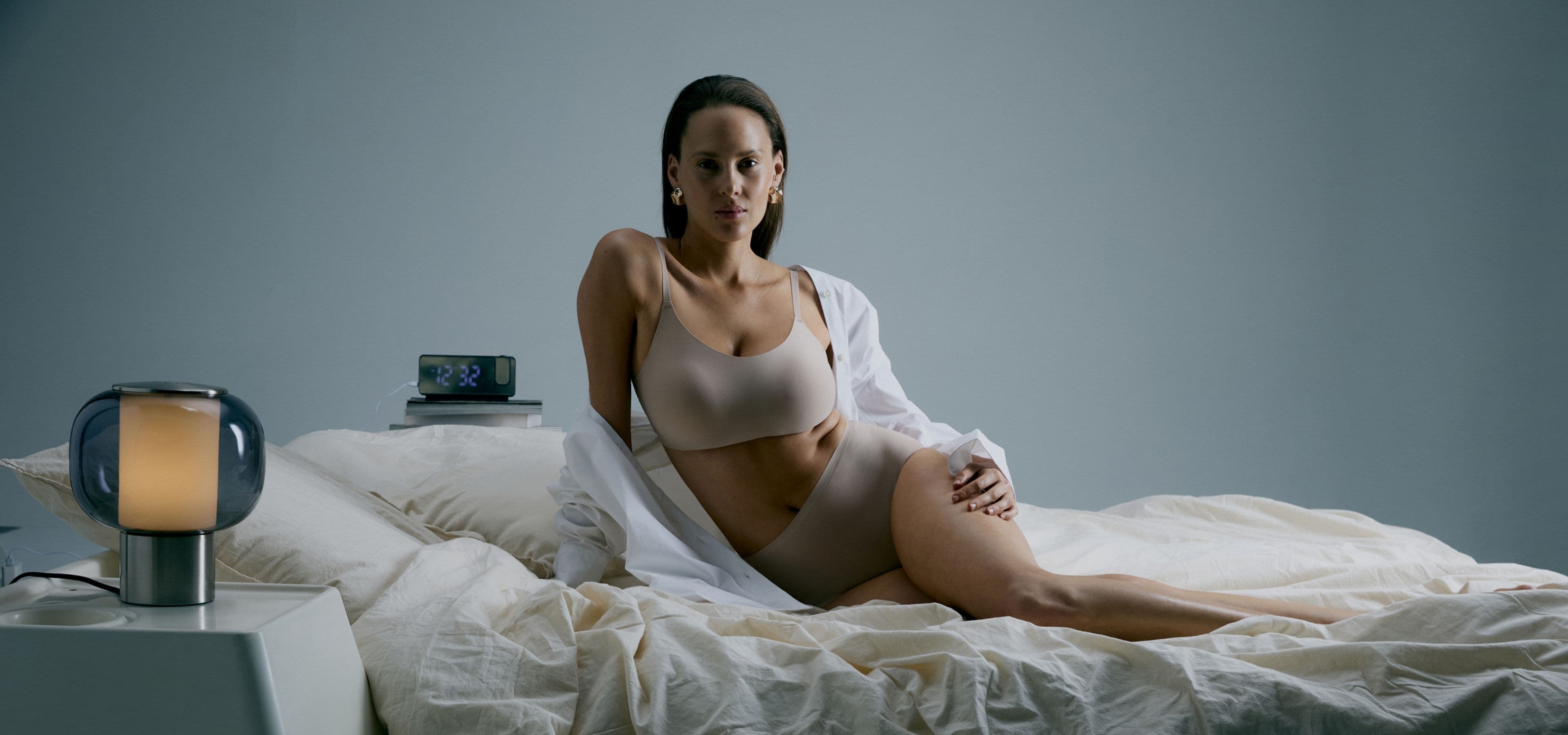 A woman in beige underwear and white shirt sits on a bed in a minimalist room, beside a lamp and digital clock showing 8:28.