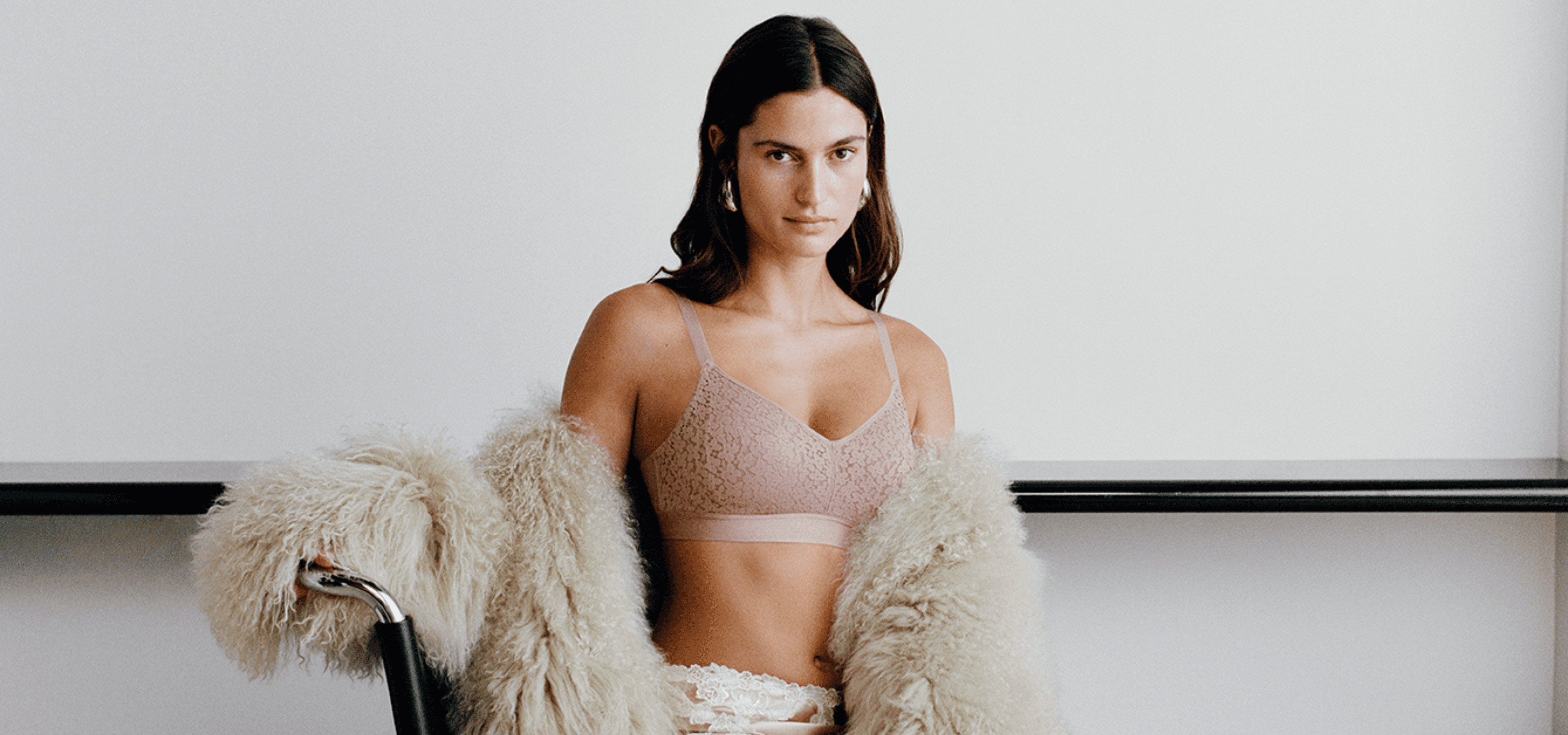 A woman in a lace bralette sits on a chair draped with a fluffy coat, against a white background.