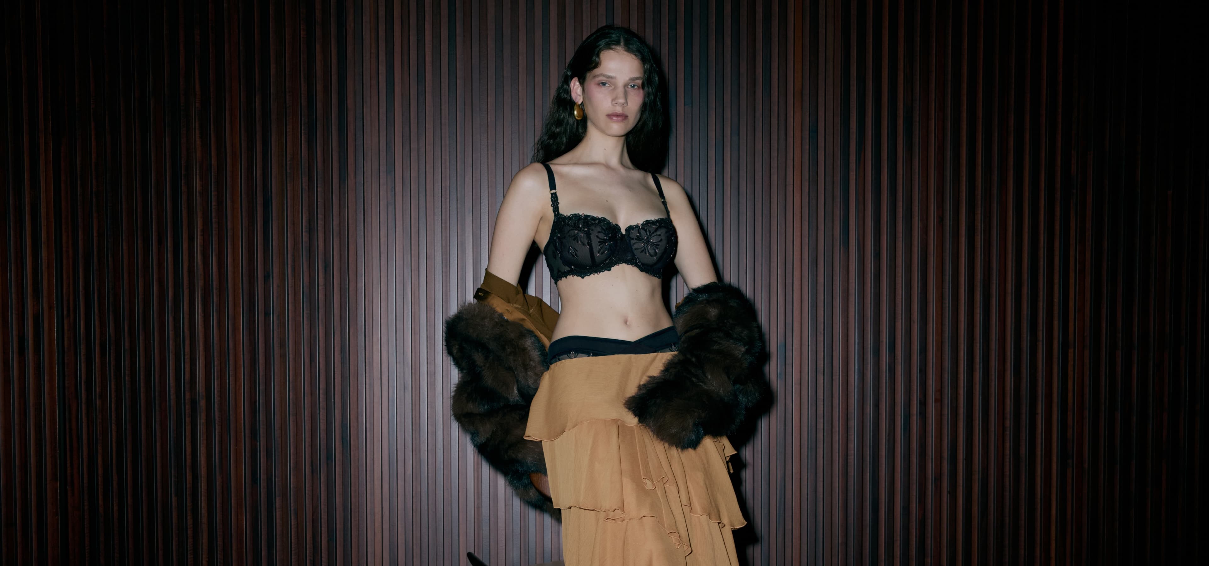 Model wearing a black lace bra, brown ruffled skirt, and a fur stole, standing against a dark wooden background.