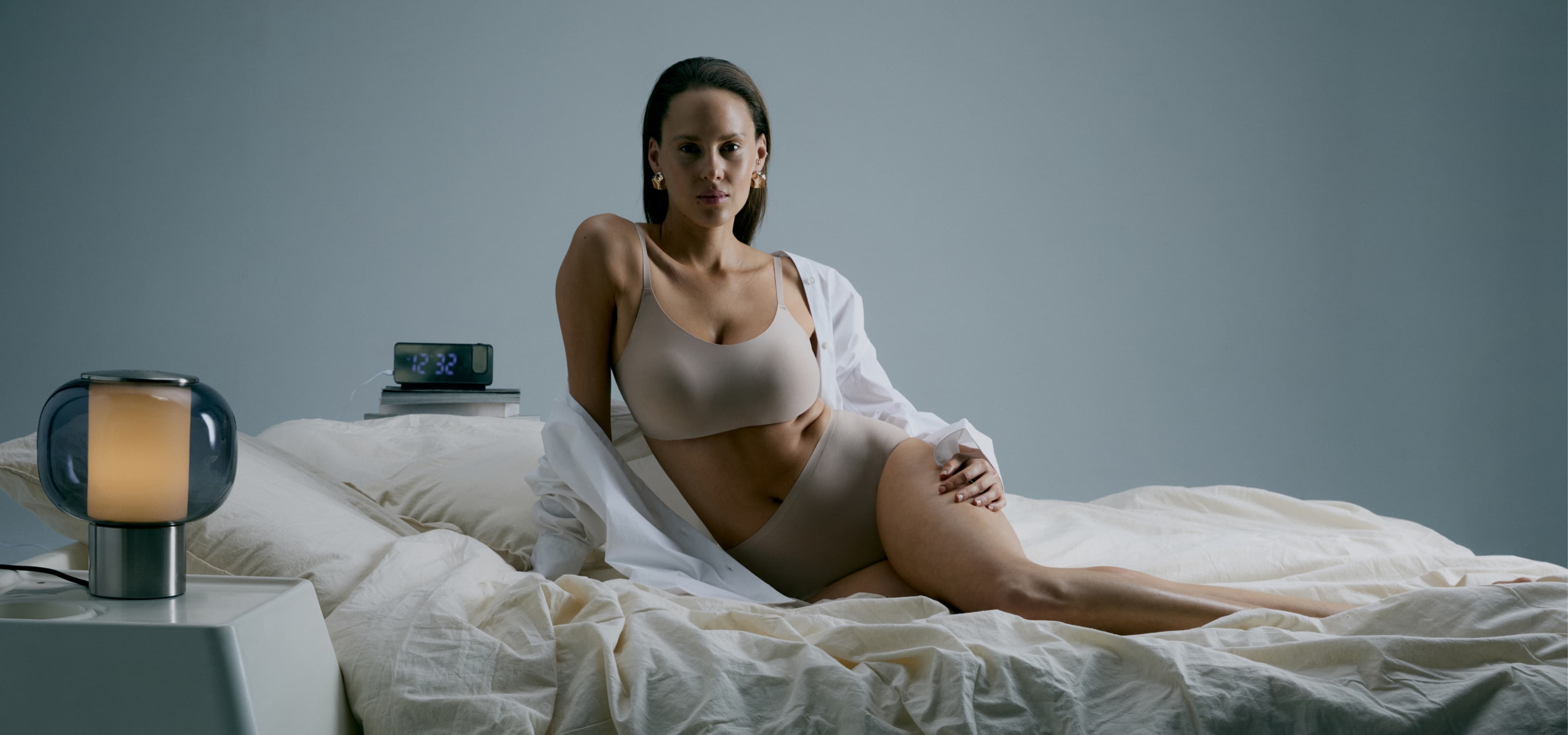 A woman in nude-toned lingerie and a white shirt lounges on a bed. A lamp and clock are on the bedside table.
