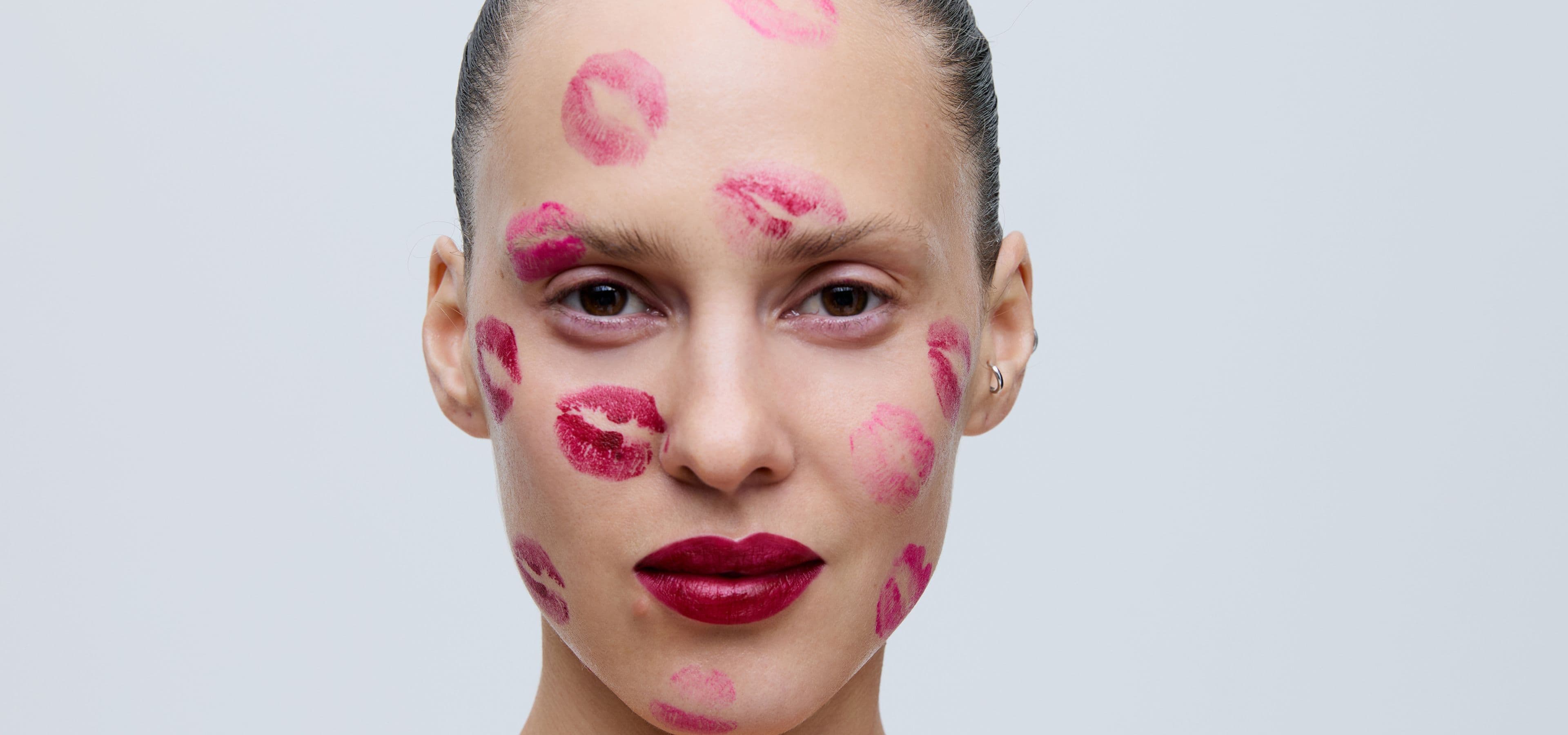 Woman with dark lipstick, covered in lipstick kiss marks on her face and forehead, against a plain background.