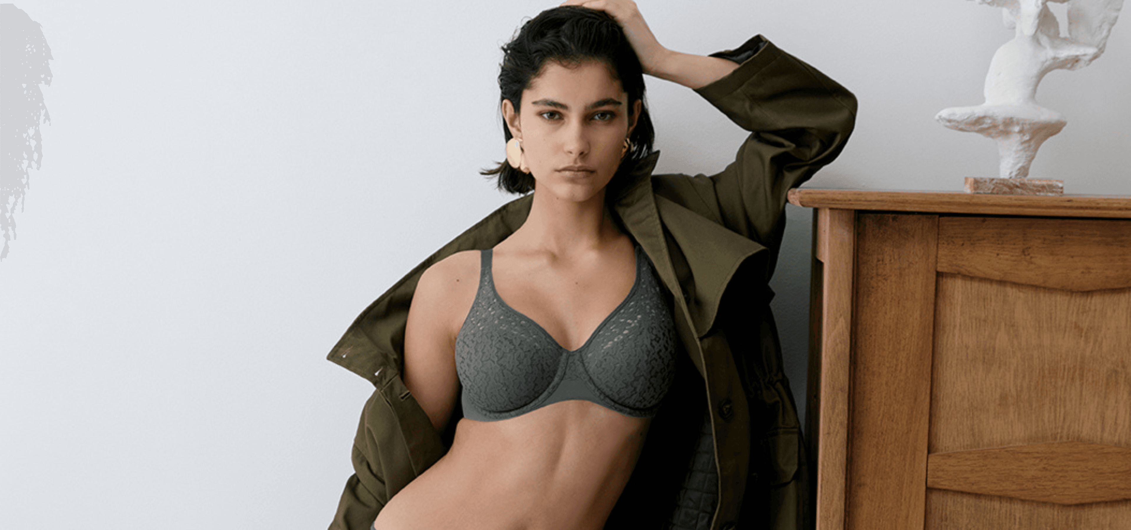 A person wearing a lace dark green bra and an open olive jacket stands confidently beside a wooden dresser with a white sculpture on it.