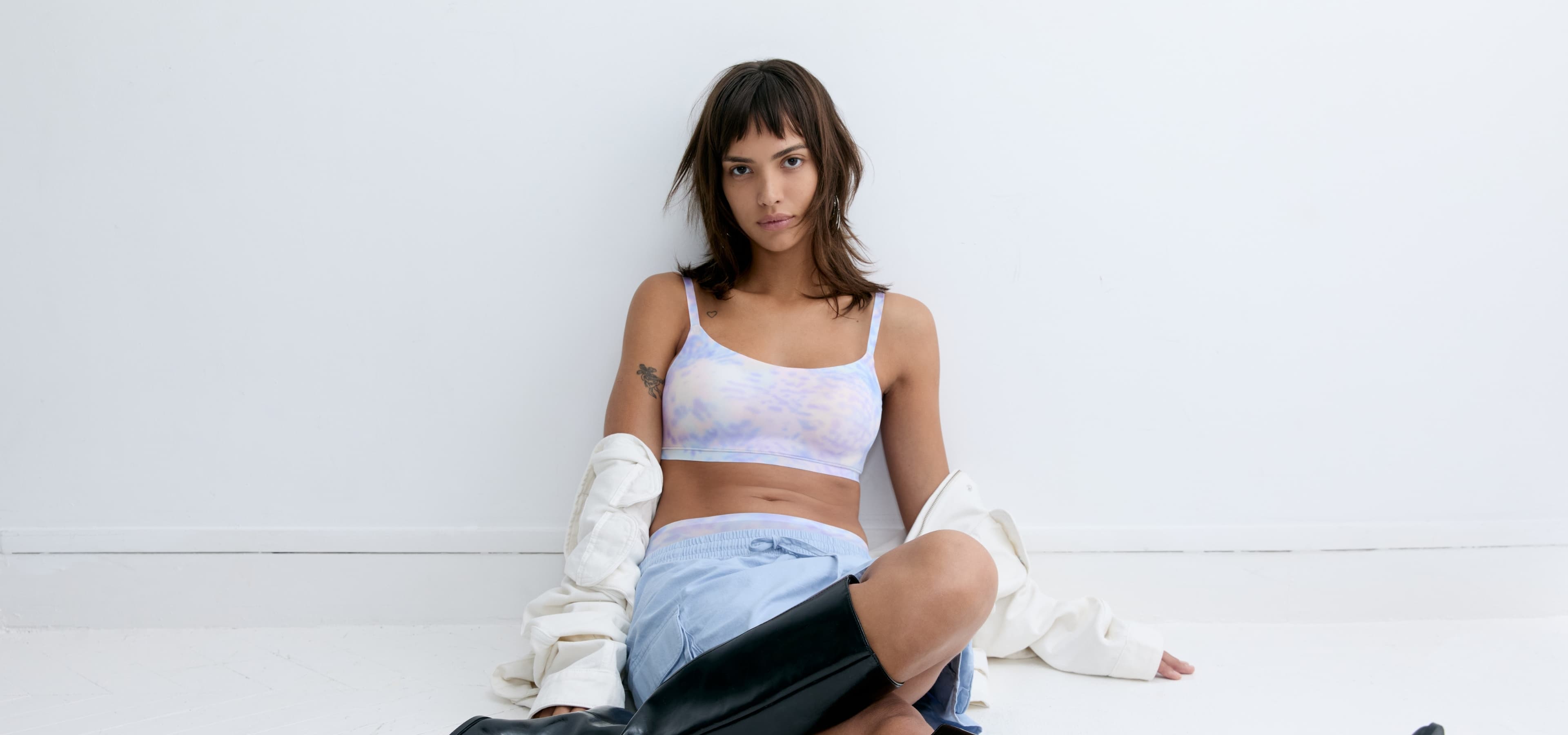 Person sitting on the floor against a white wall, wearing a  pastelle bra, blue shorts, black boots, and holding a white jacket.