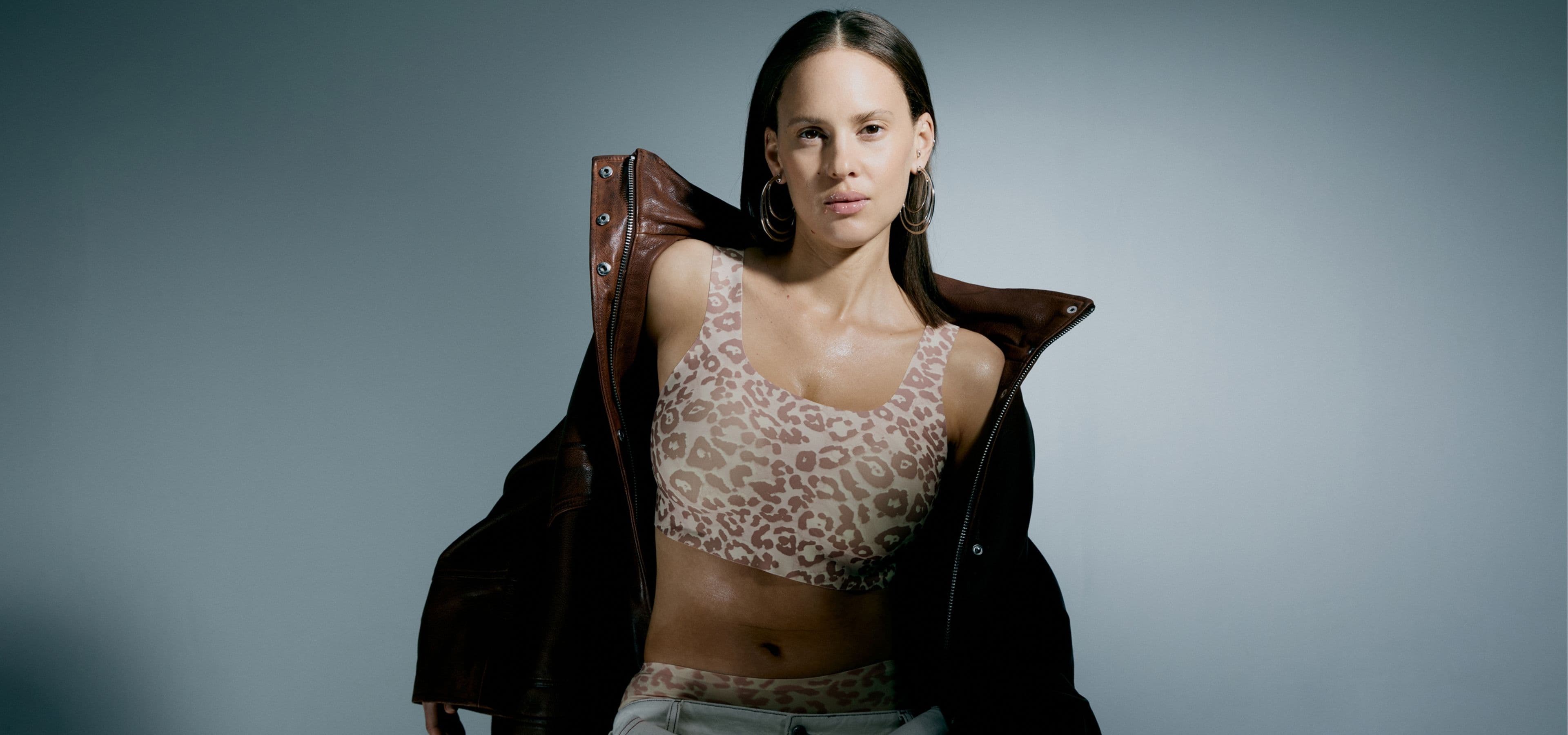 A woman in a leopard print top and brown jacket poses confidently against a gray background.