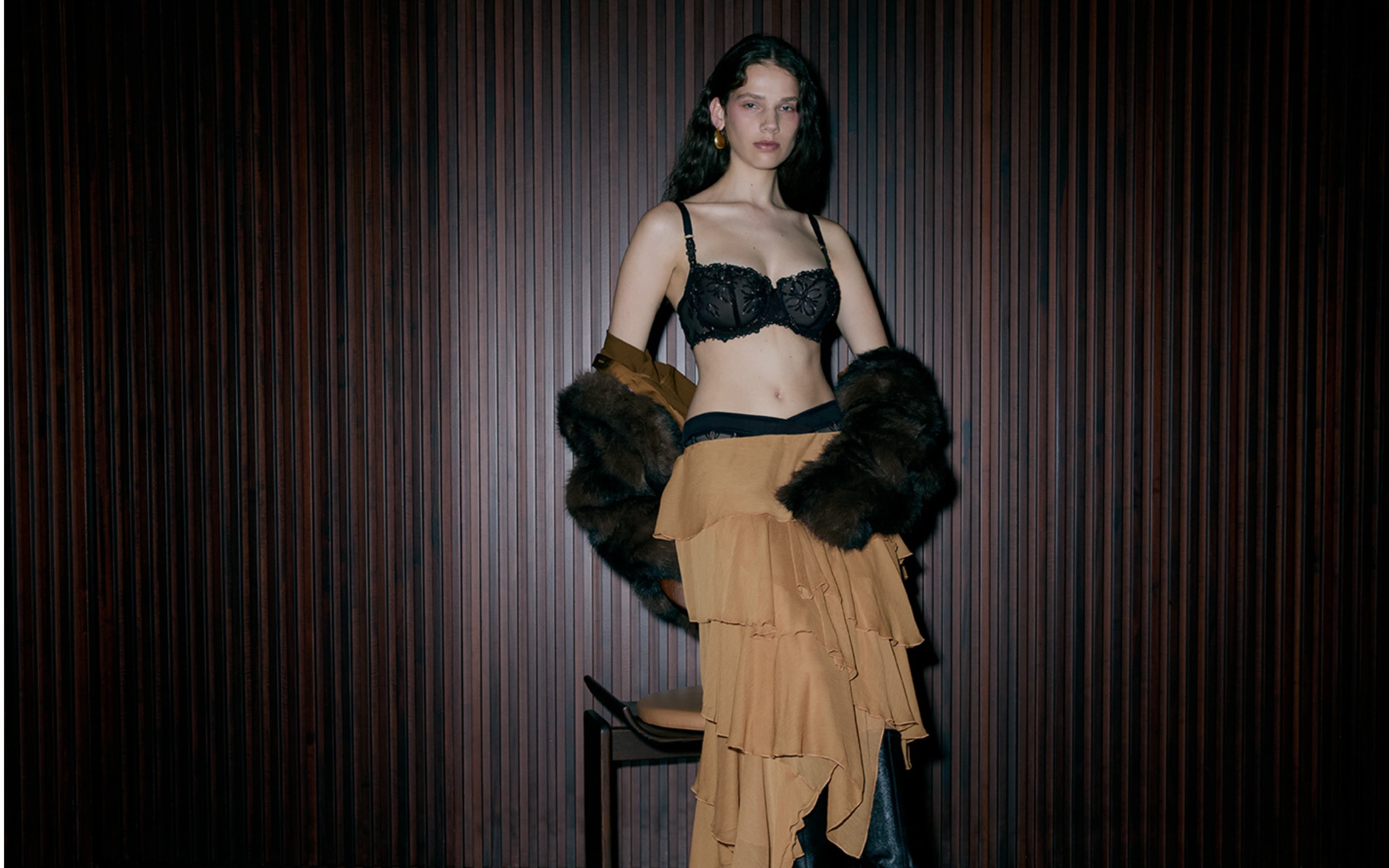 A woman in a black lace bra and layered tan skirt stands against a dark, striped wall, draped in a fur coat and wearing gold earrings.