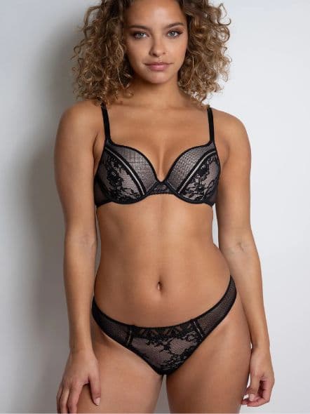 Person wearing black lace lingerie, standing against a plain white background, looking confidently at the camera.