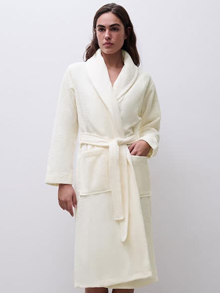 Person wearing a cream-colored bathrobe with a shawl collar and belt, standing against a plain white background.