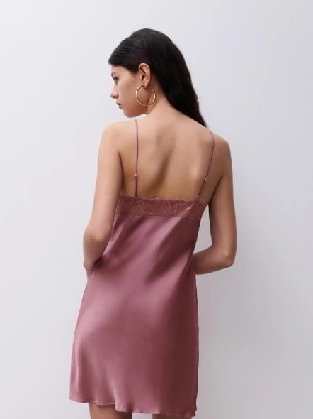 Woman in silk pink slip dress with lace detail, large hoop earrings, facing a white background.