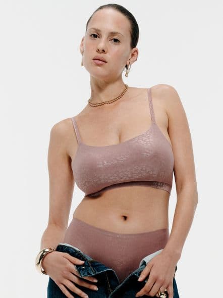 Woman in mauve seamless bralette and high‑waist underwear with low jeans, wearing gold hoop earrings and necklace against a neutral background.