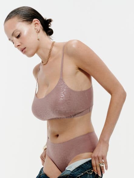 Woman in a mauve leopard-print lingerie set, with slicked-back hair, adjusts her jeans. She wears gold jewelry against a plain white background.