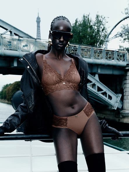 Person in brown lingerie and black jacket poses confidently on a boat, with a bridge and the Eiffel Tower in the background.