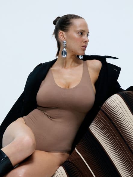 Person in a tan bodysuit, black coat, and large earrings sits on a patterned chair, gazing to the side against a plain background.