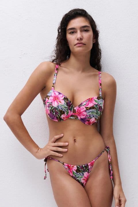 Woman in a floral bikini stands confidently against a plain white background, with long curly hair and arms relaxed by her sides.