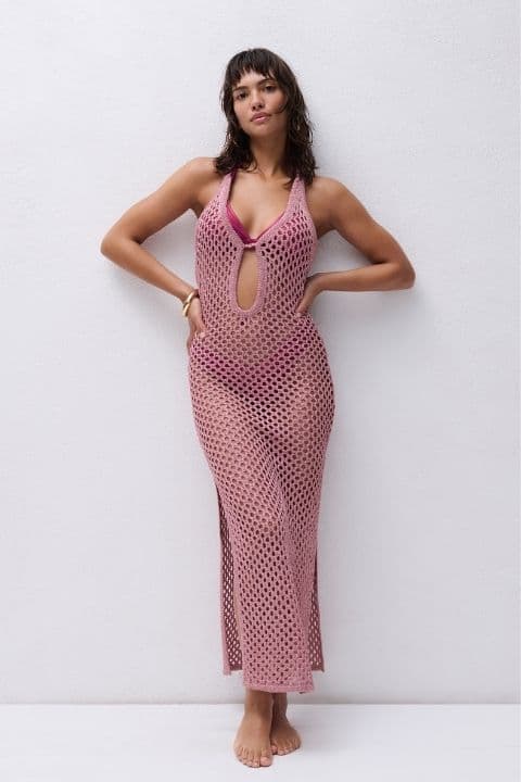 Woman in a pink crochet dress with a keyhole design stands barefoot against a white wall, looking confidently at the camera.