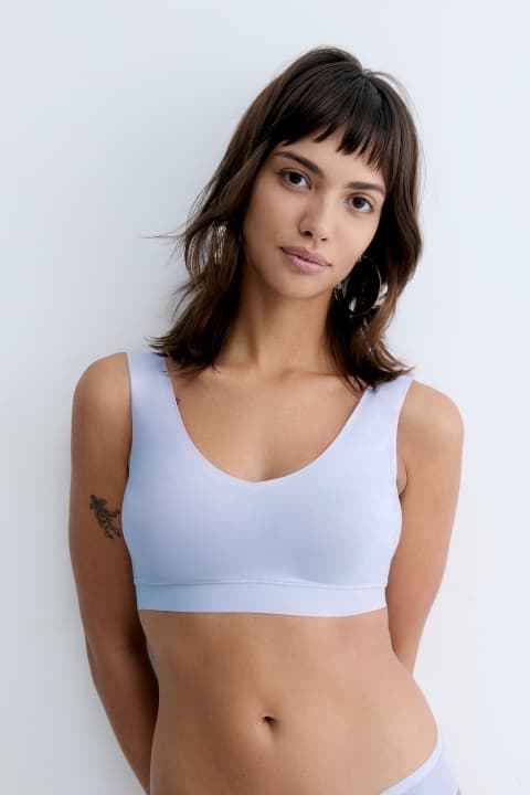 A woman with shoulder-length hair, wearing a light blue sprts bra, poses confidently against a white background. She has a tattoo on her upper arm.