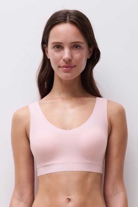 Woman in a light pink bra, standing against a plain white background, with a neutral expression.
