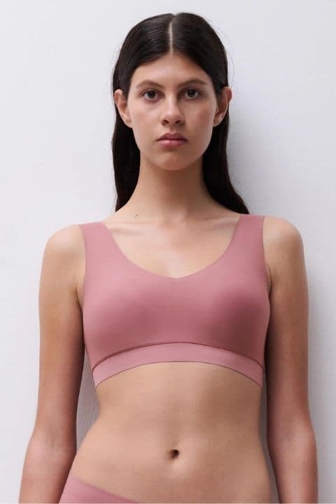 A woman with long dark hair wears a pink sports bra, standing against a plain white background, looking directly at the camera.