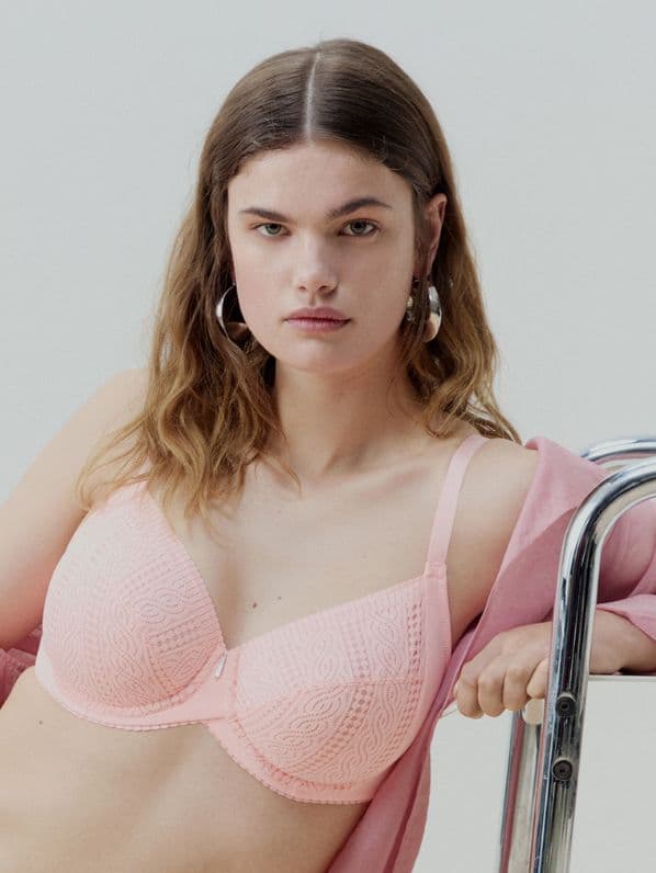 Woman wearing a pastel pink lace bra and silver hoop earrings, leaning on a metal rail, with a soft expression.