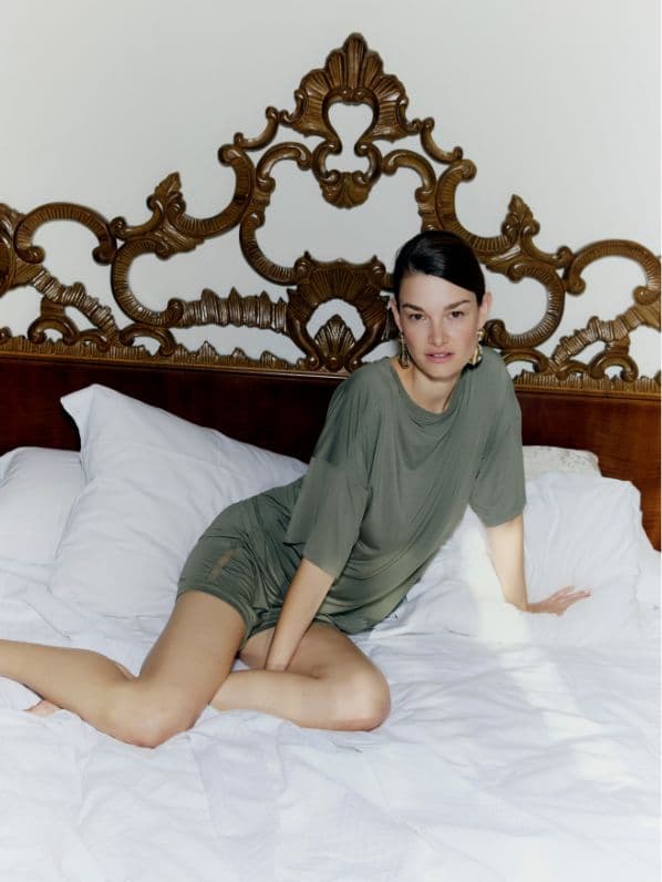 A person in a loose green dress poses on a bed with ornate, gold headboard, surrounded by white pillows and bedding.