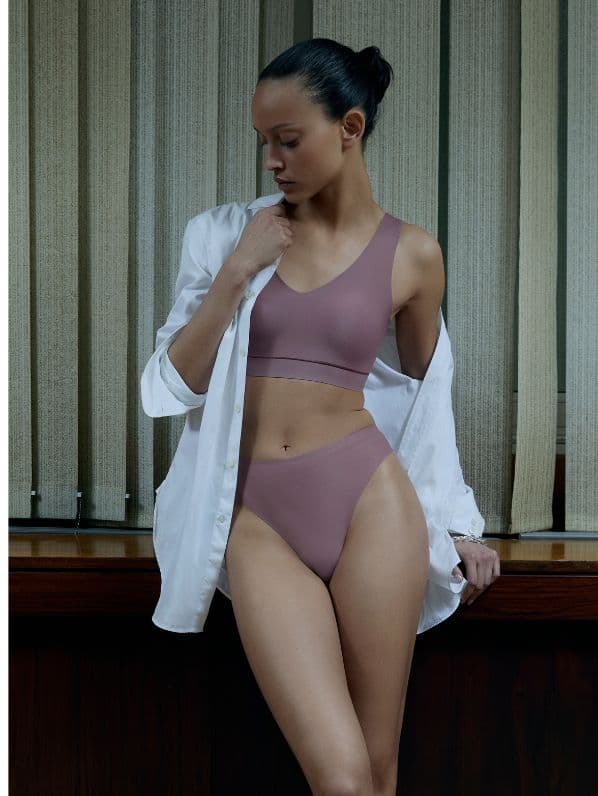 A woman in mauve underwear and a white shirt leans against a wooden ledge, with vertical blinds in the background.