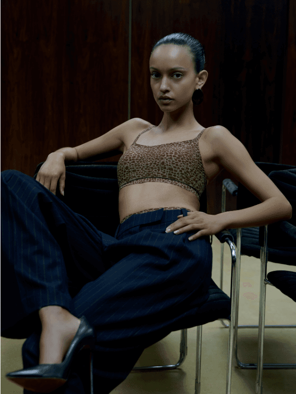 A woman in a leopard print crop top and pinstripe pants sits confidently on a chair with a wooden background.