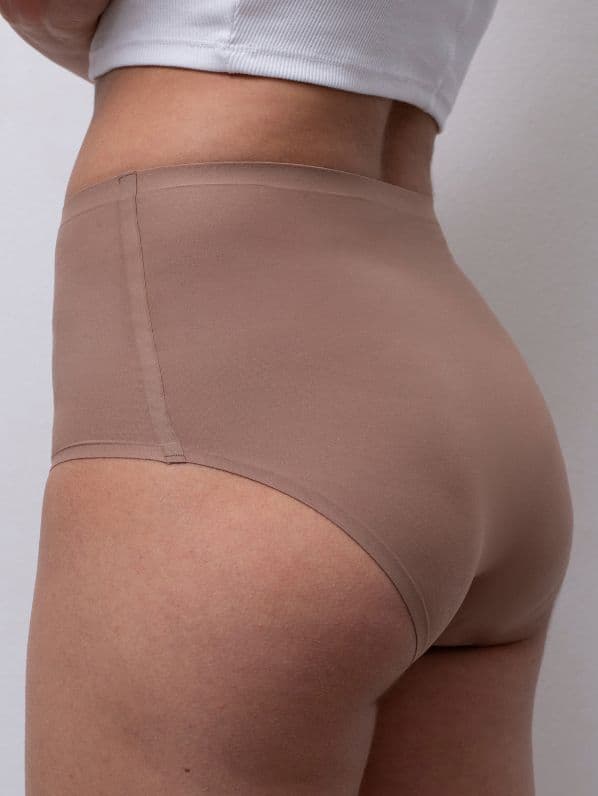 Person wearing seamless, tan-colored high-waisted underwear, standing sideways against a white wall.