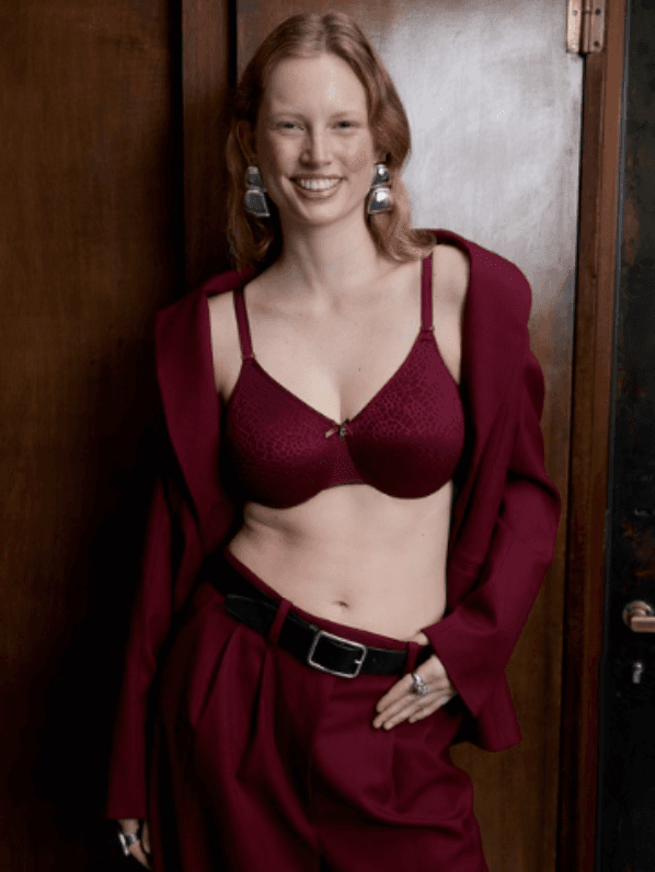 A woman smiles wearing a burgundy bra and matching tailored pants with a jacket draped over her shoulders, standing against a wooden door.