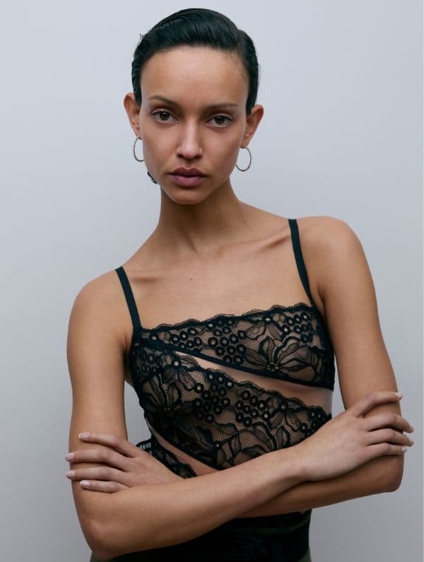 Person with short hair wearing a black lace top, arms crossed, and hoop earrings, against a plain background.