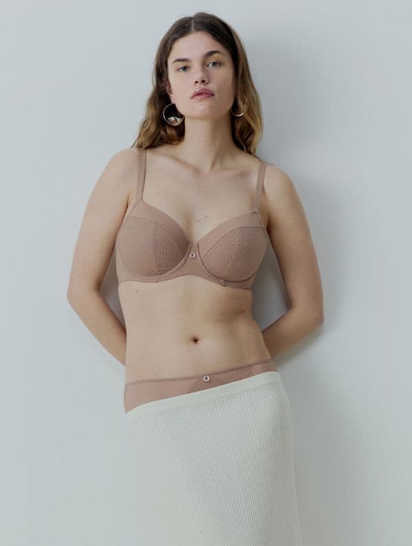 Woman wearing a beige bra and cream skirt stands against a light gray background, with long hair and hoop earrings.